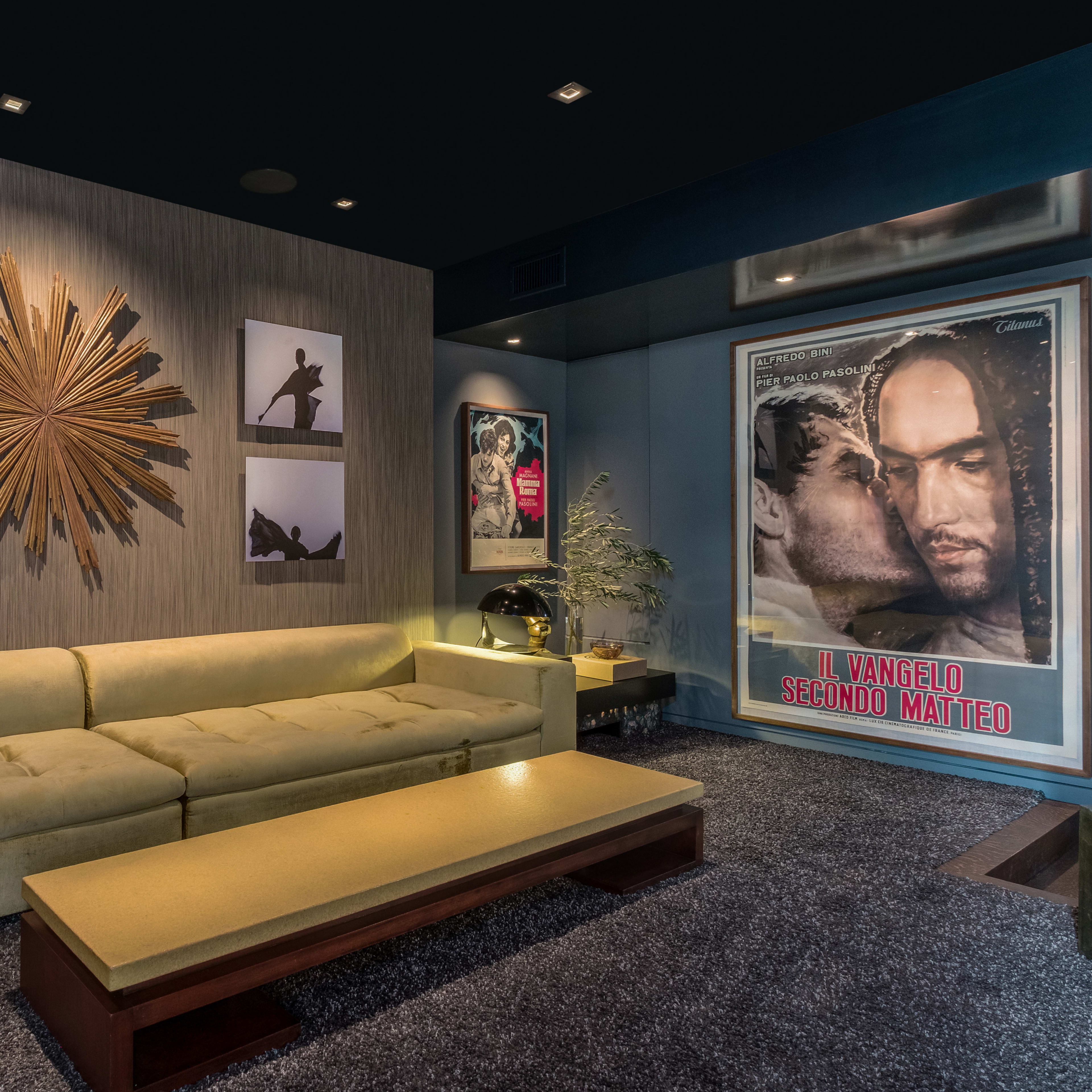 A modern living room featuring a yellow sectional couch, a coffee table, framed artwork on the walls, and a large movie poster.