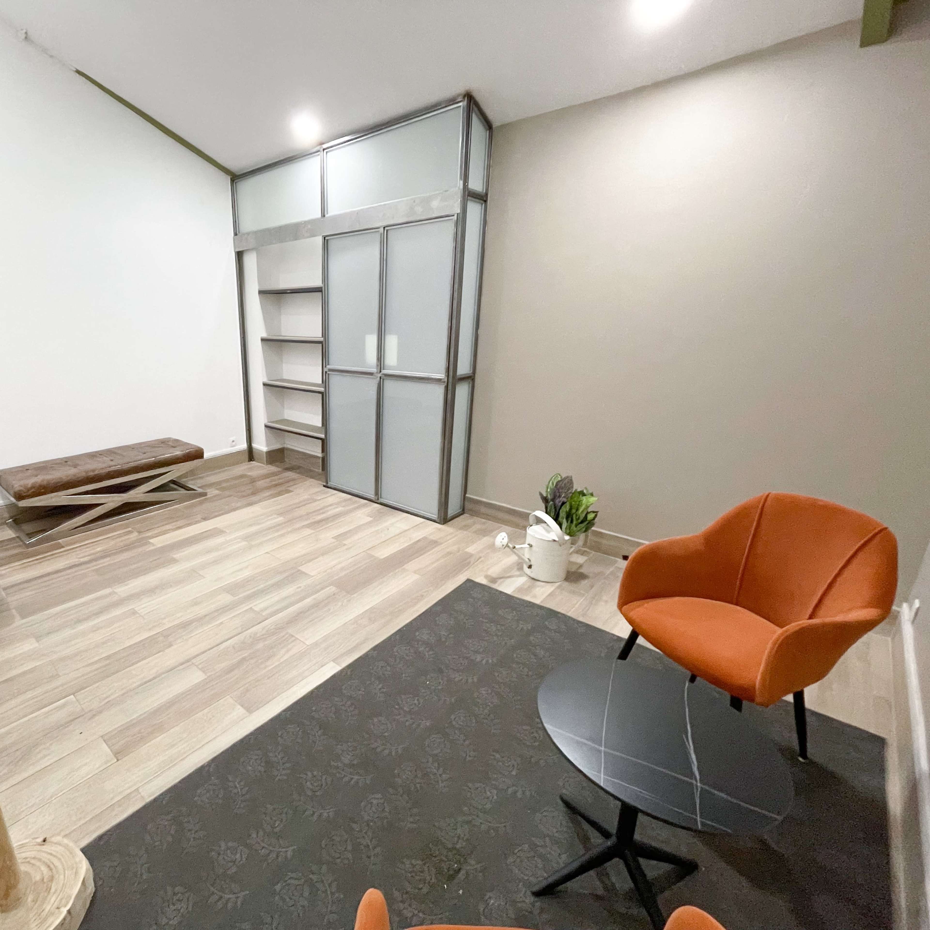 A minimalist interior space featuring two orange chairs, a small round black table, a wooden coat rack, and a storage unit against the wall.