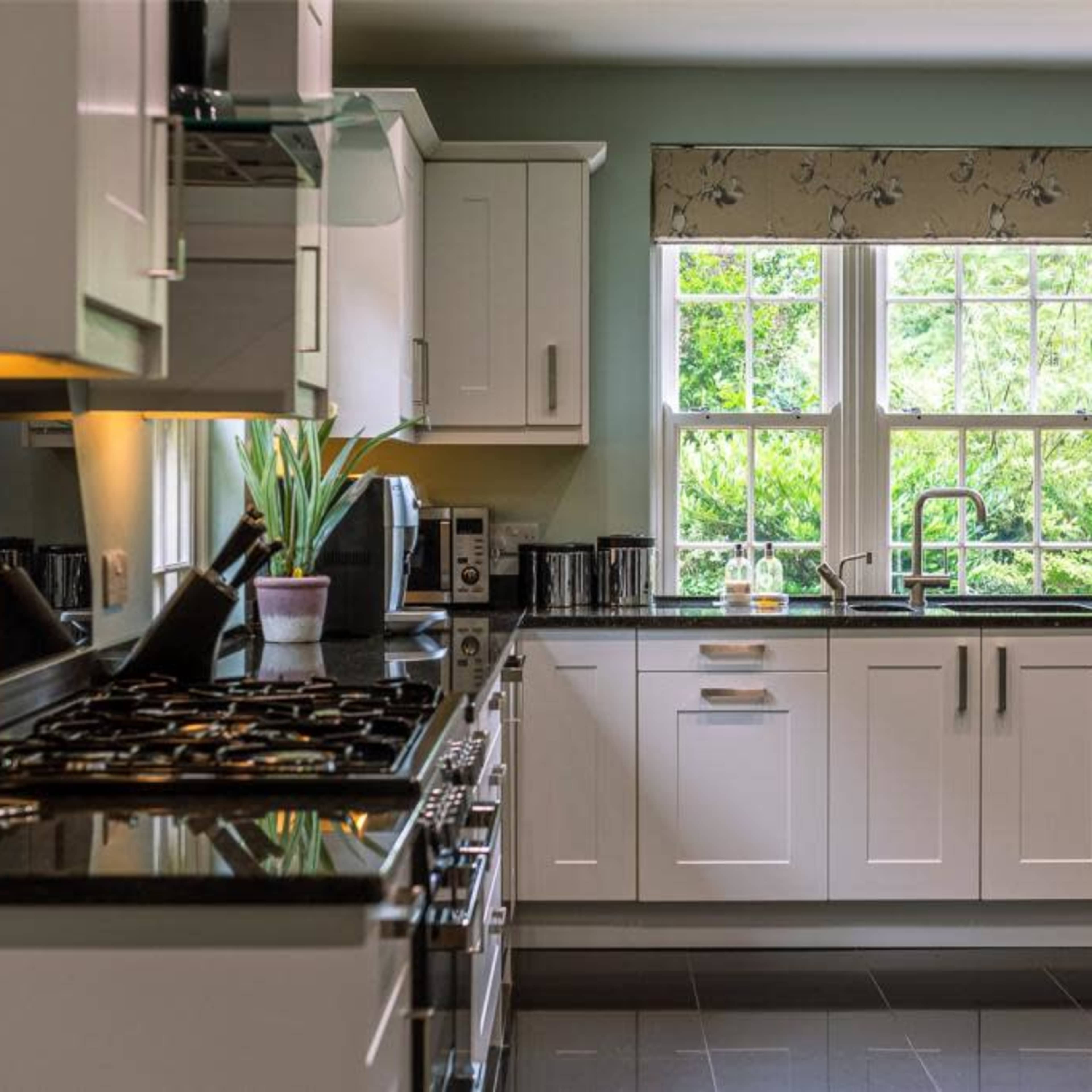 A modern kitchen features white cabinetry, a black countertop, and large windows overlooking greenery outside.