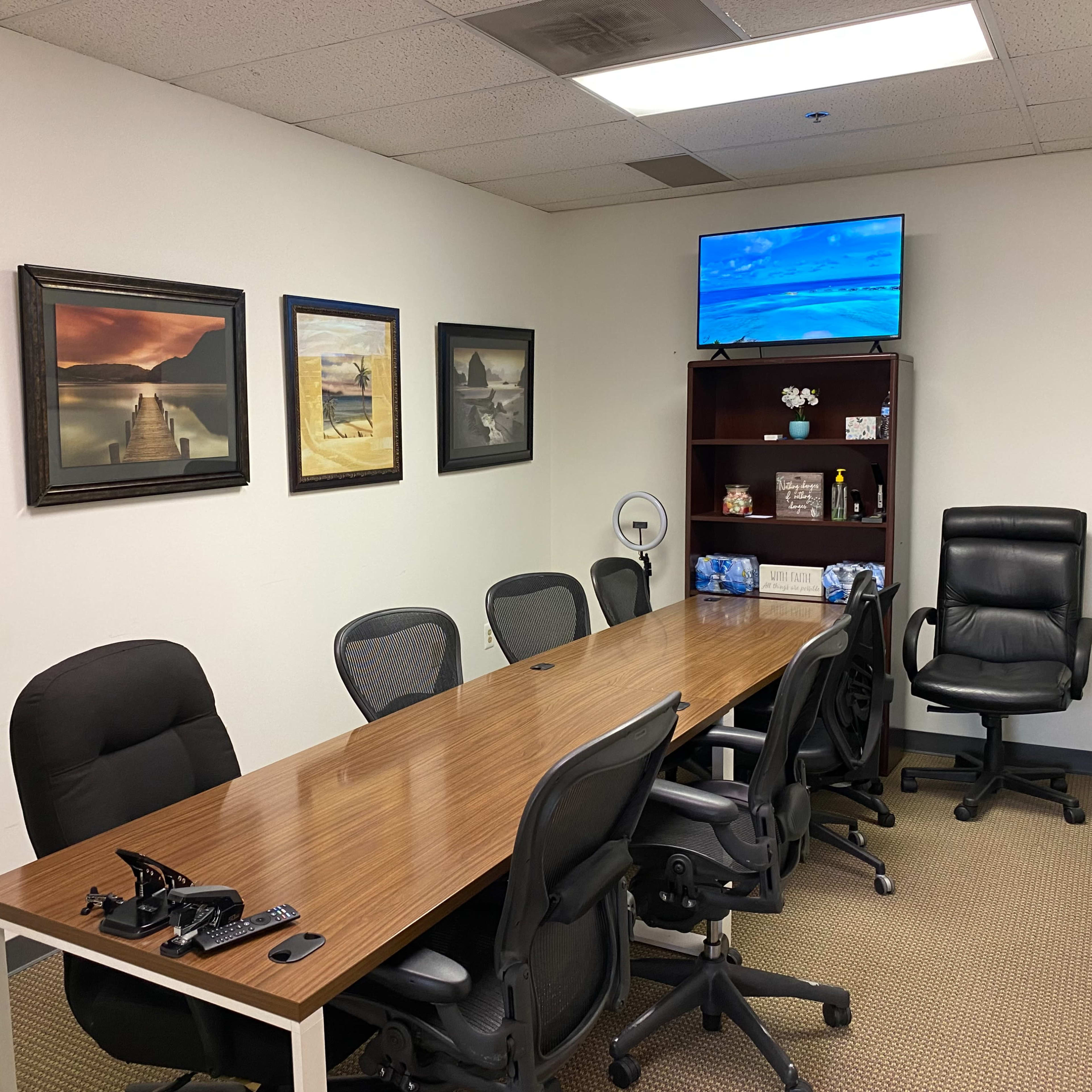 A conference room with a long wooden table, several office chairs, framed pictures on the walls, and a television mounted in one corner.