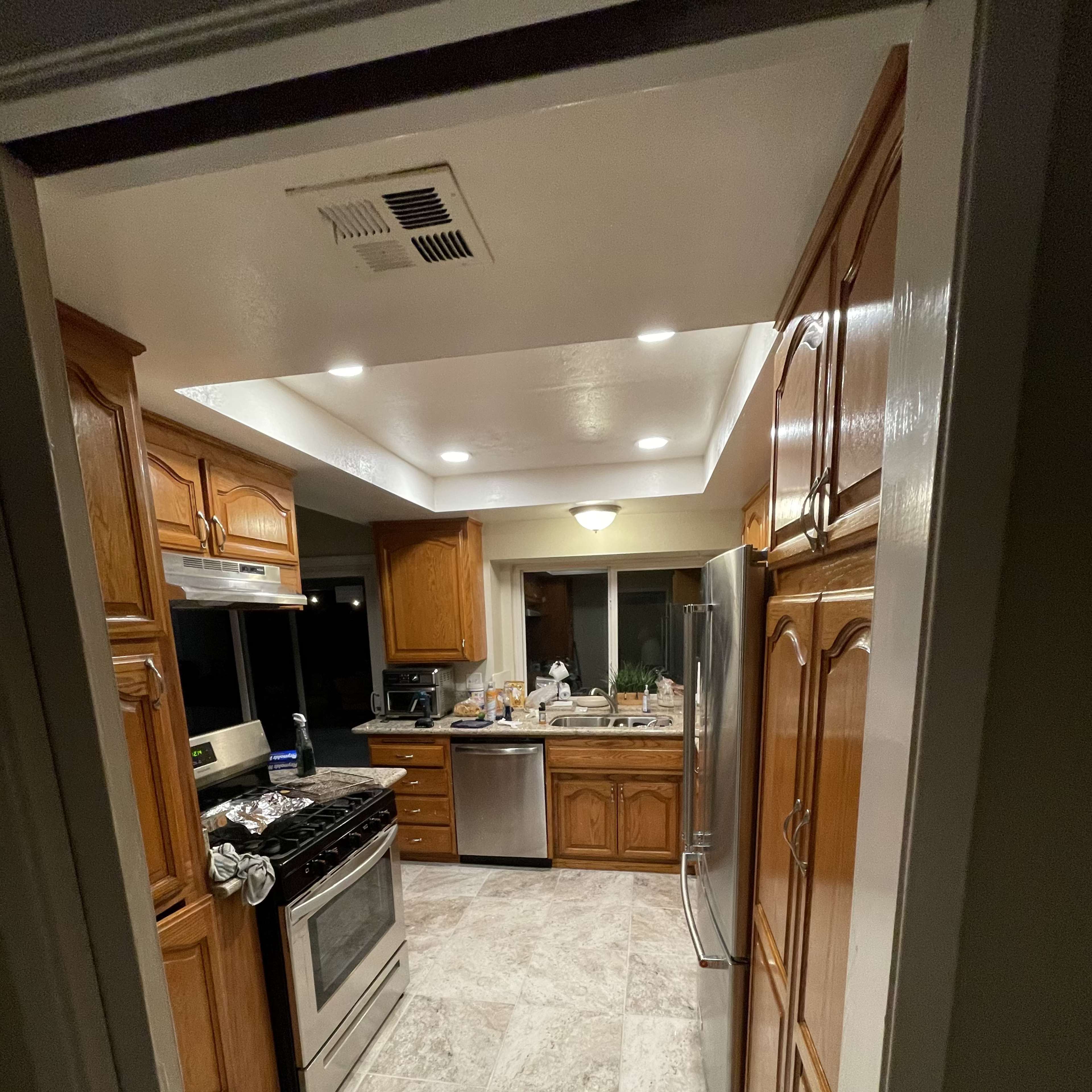 A kitchen with wooden cabinets, a stainless steel refrigerator and stove, and a tiled floor, viewed from an adjacent hallway.
