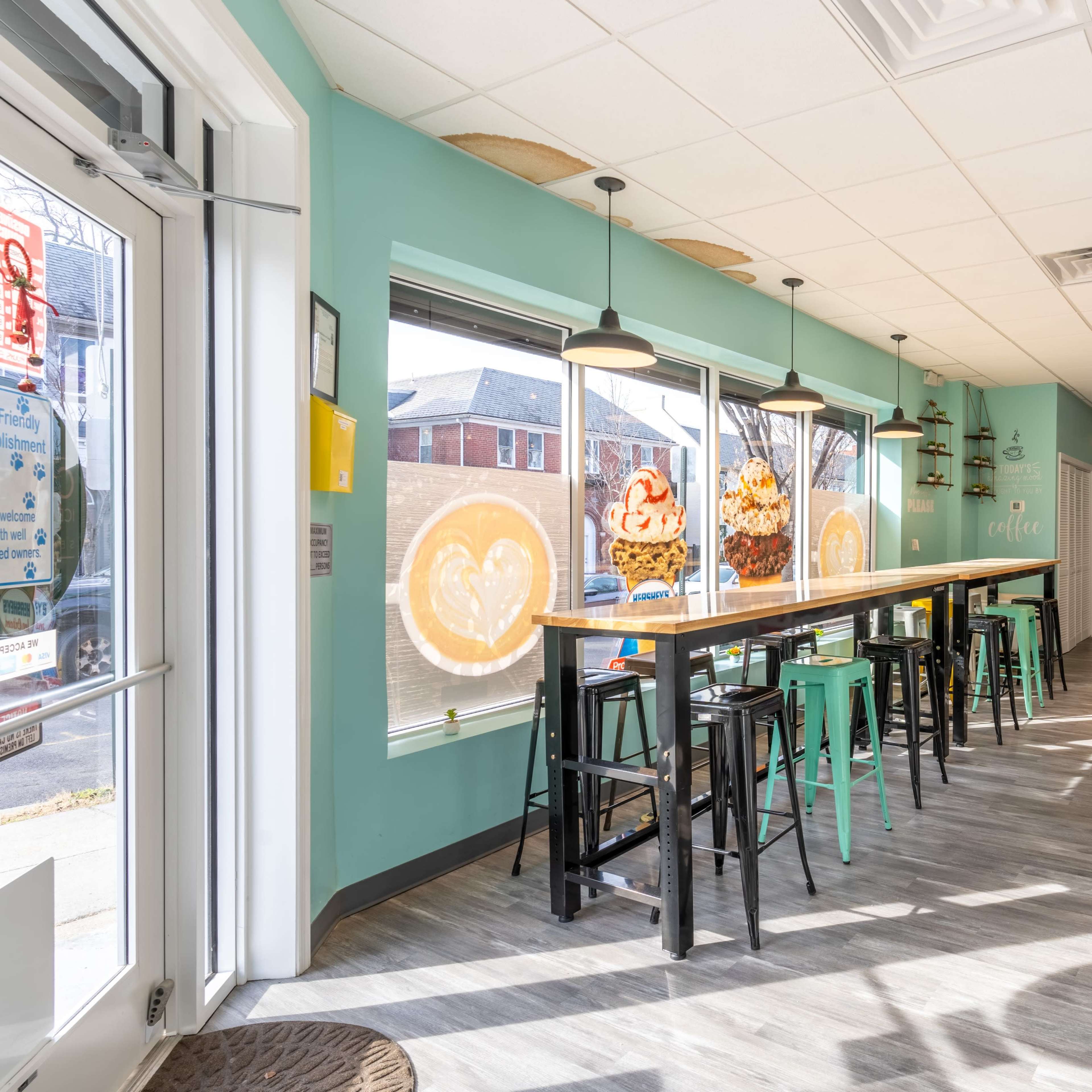 A brightly lit café interior with tall tables and stools, large windows adorned with colorful artwork, and a retail area displaying various snacks and drinks.