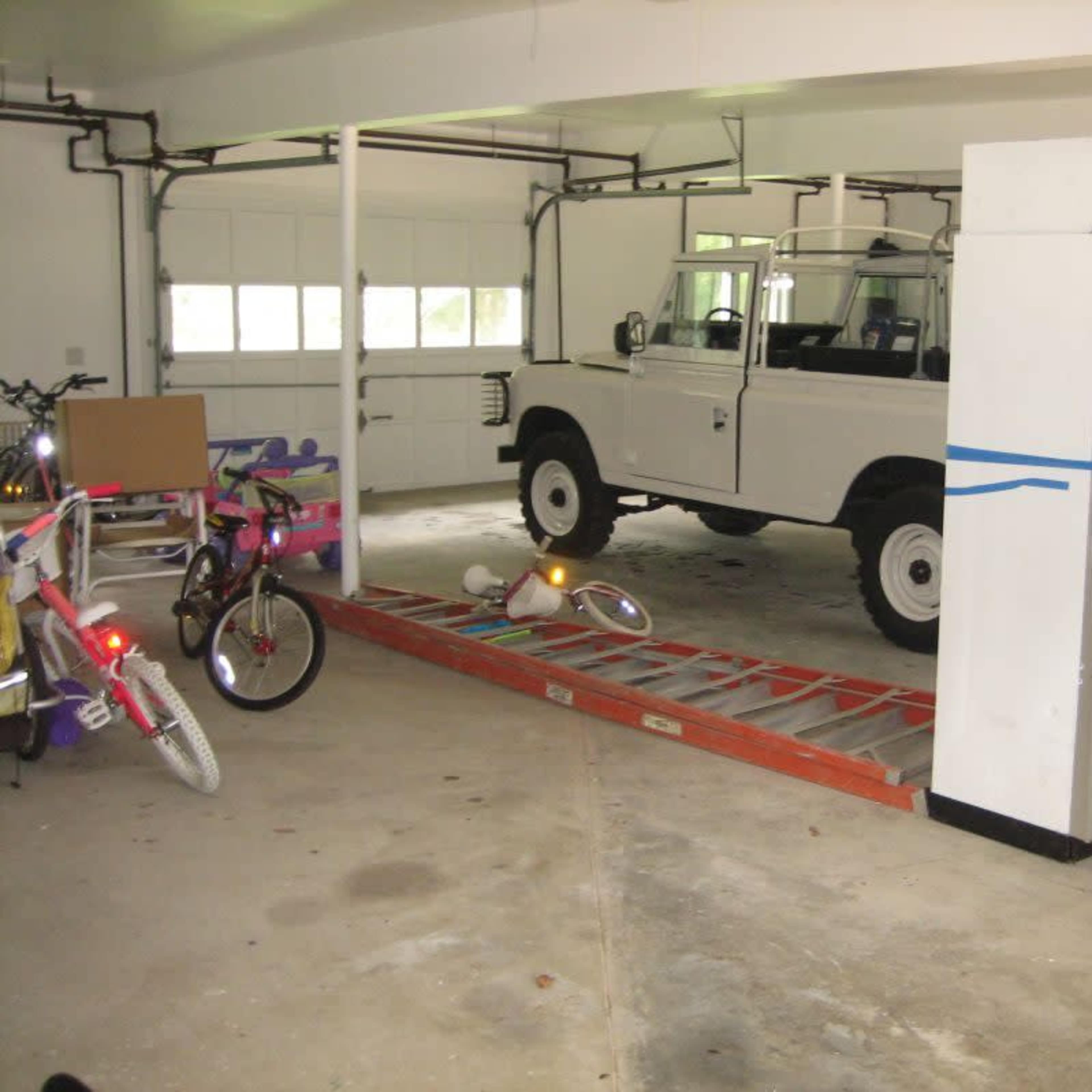 A white vehicle is parked in a spacious garage alongside various bicycles and a ladder.