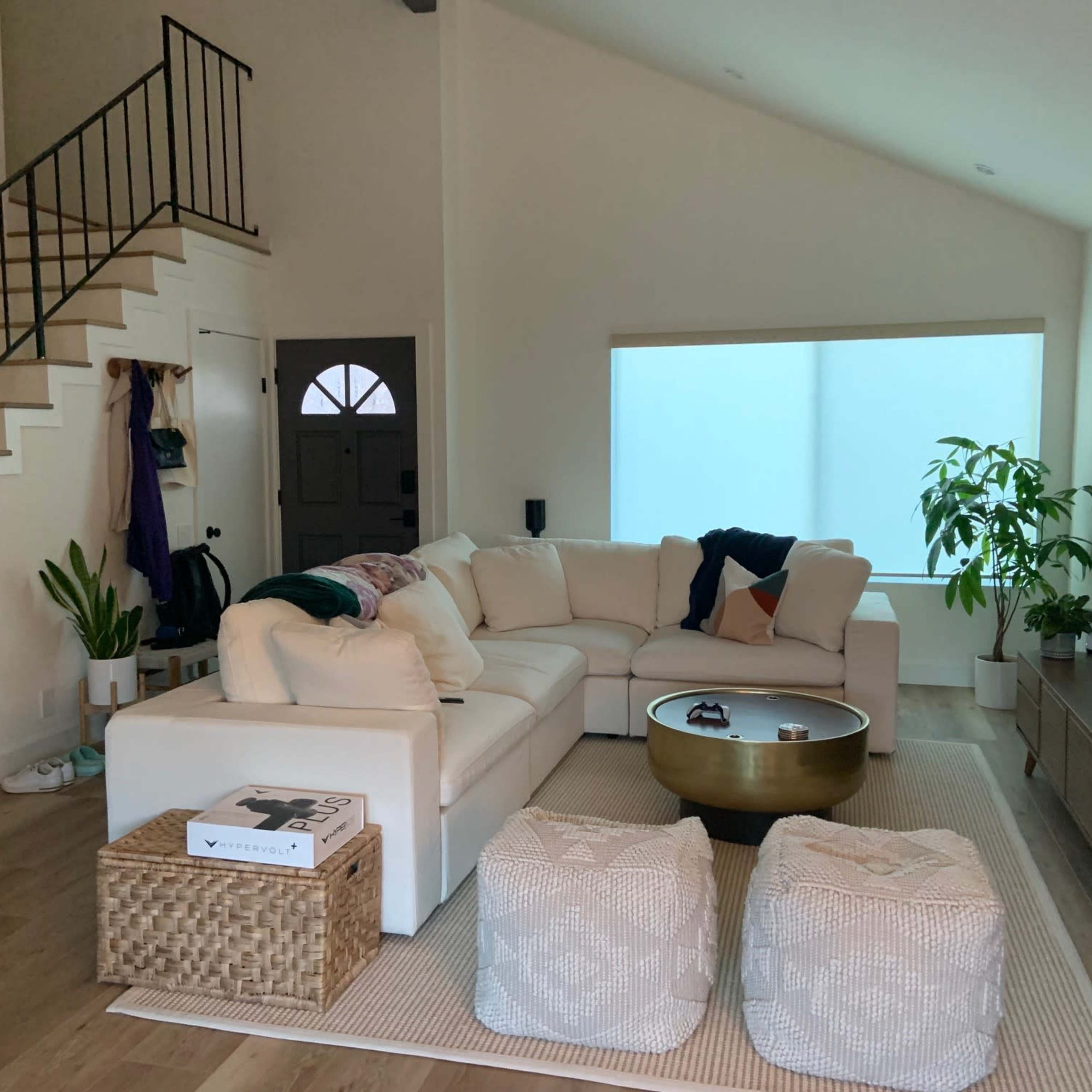A light-colored sectional sofa is arranged in a living room with a staircase, a large window covered by sheer curtains, and decorative plants.