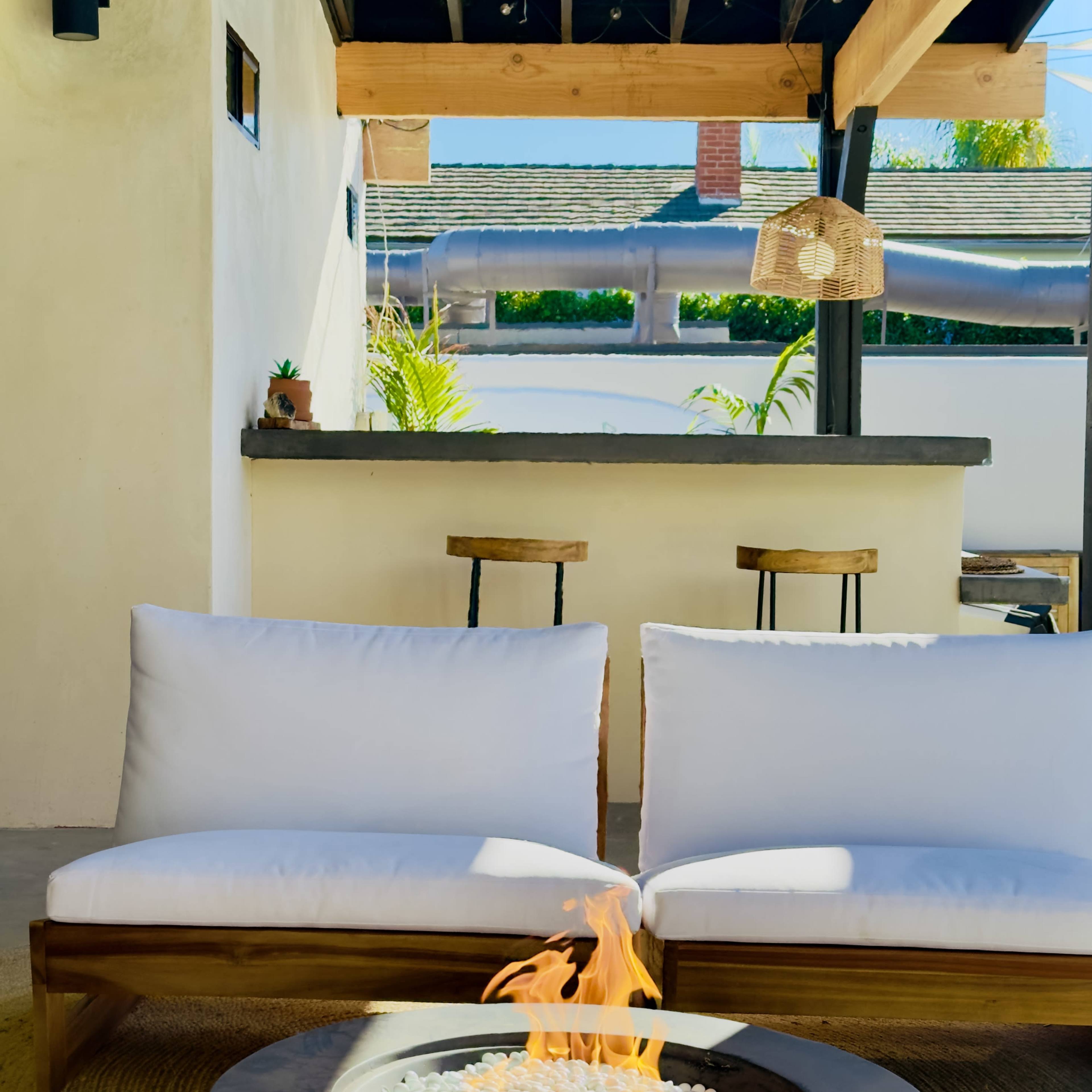 A cozy outdoor seating area featuring two white cushioned chairs facing a round fire pit, with a bar area and greenery in the background.