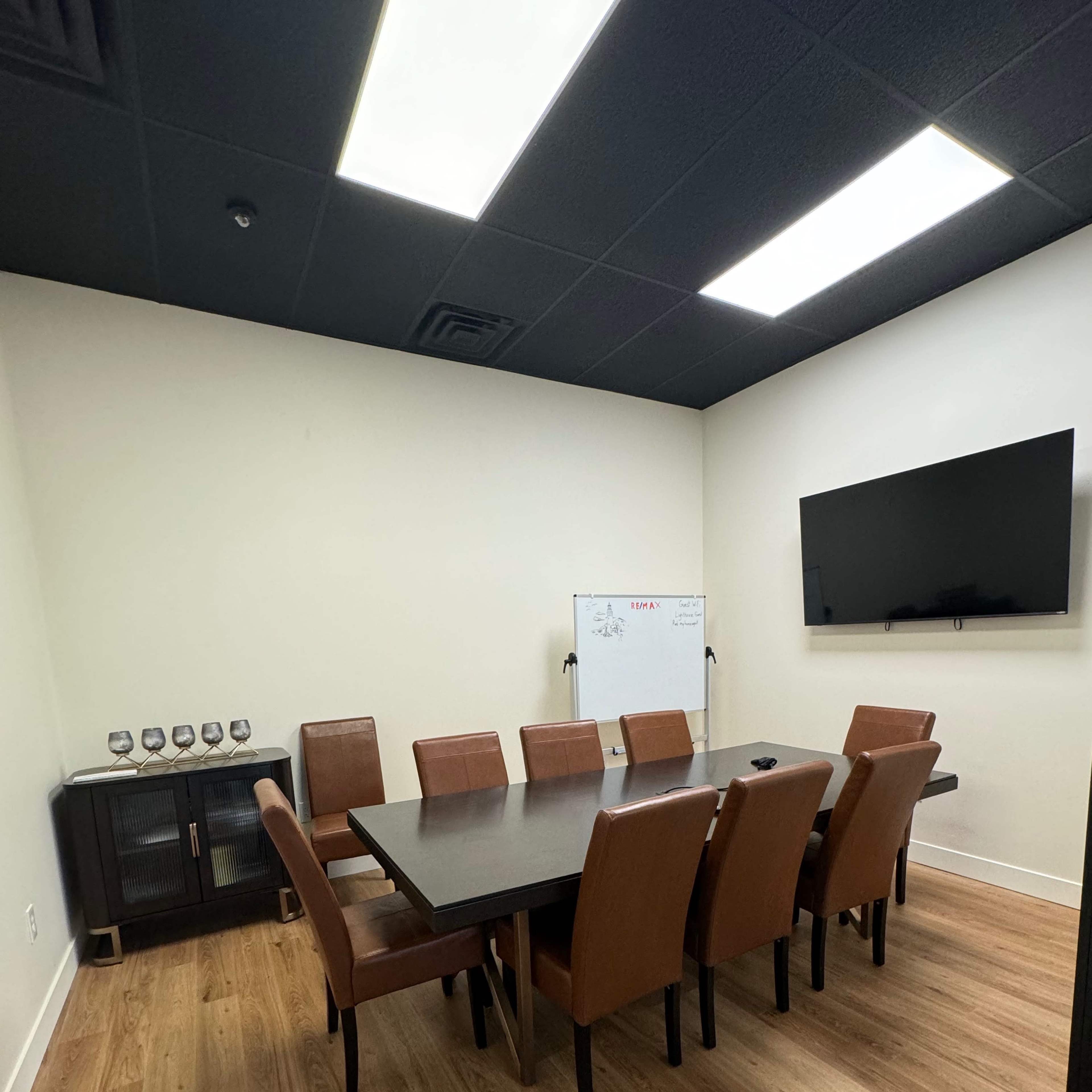A modern conference room with a large rectangular table surrounded by eight brown chairs, a wall-mounted television, and a whiteboard in the corner.