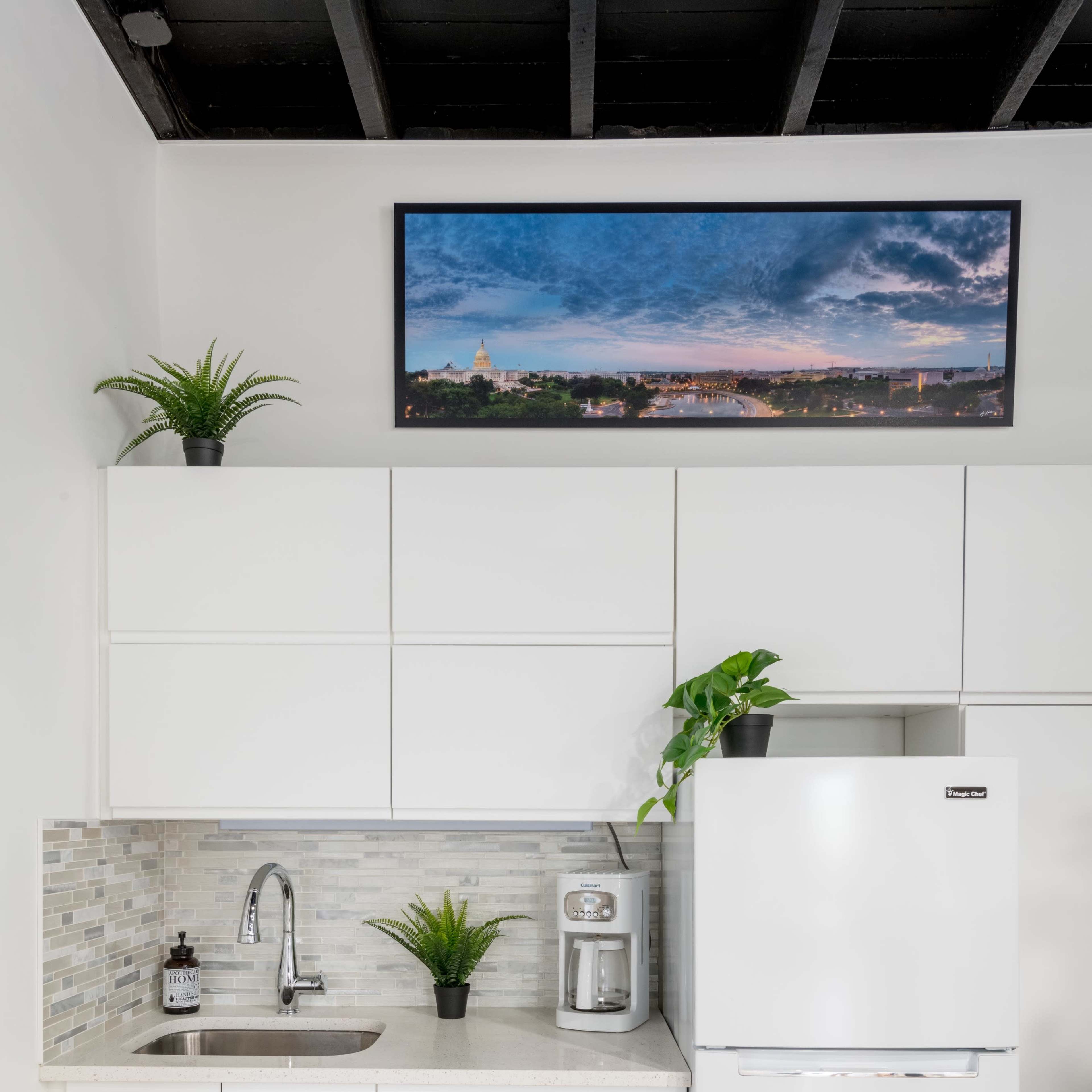 A modern kitchen setup with white cabinets, a sink, a coffee maker, and a white refrigerator, under a framed photo of a landscape.