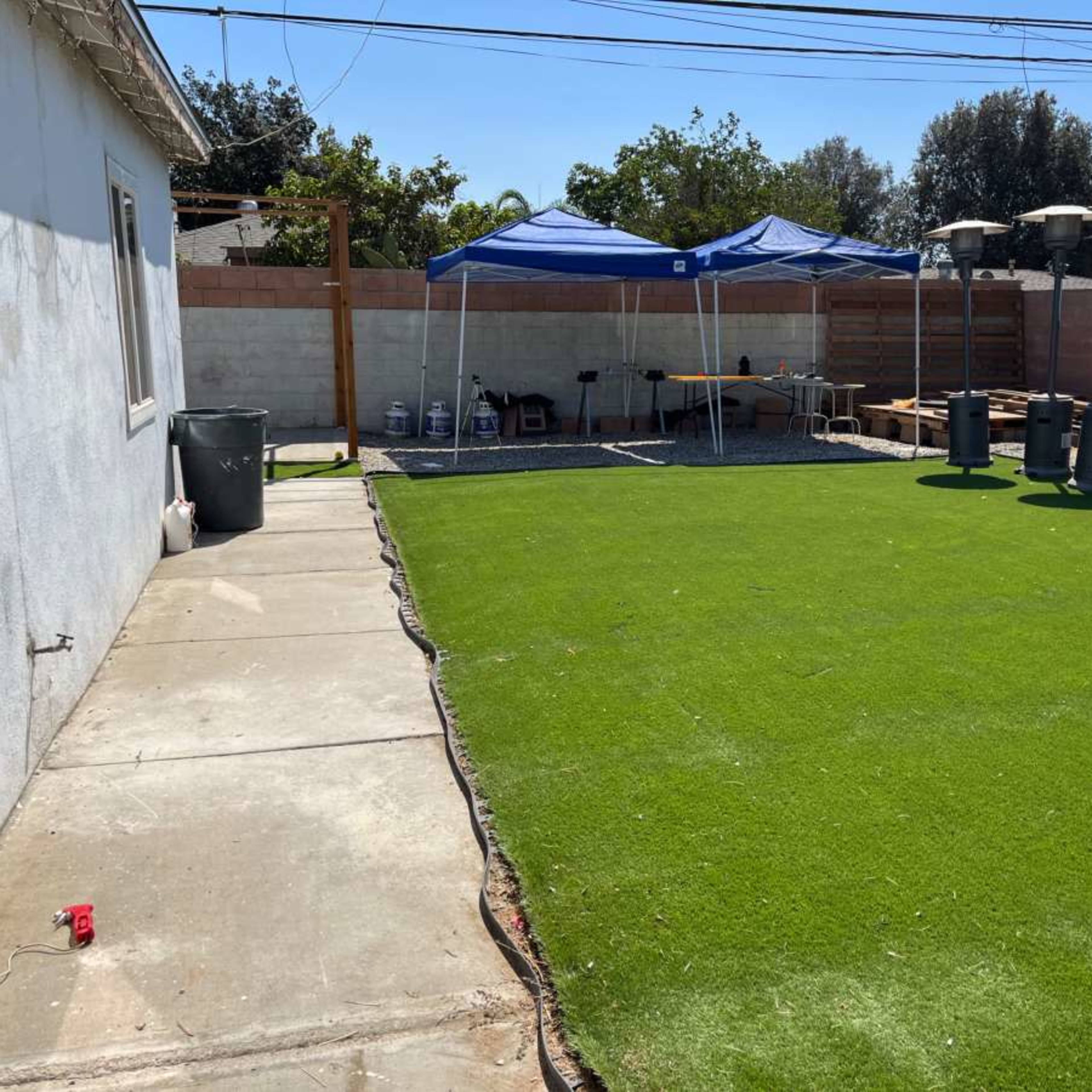 A backyard with a green artificial lawn, two blue tents set up, and several patio heaters along a fence.