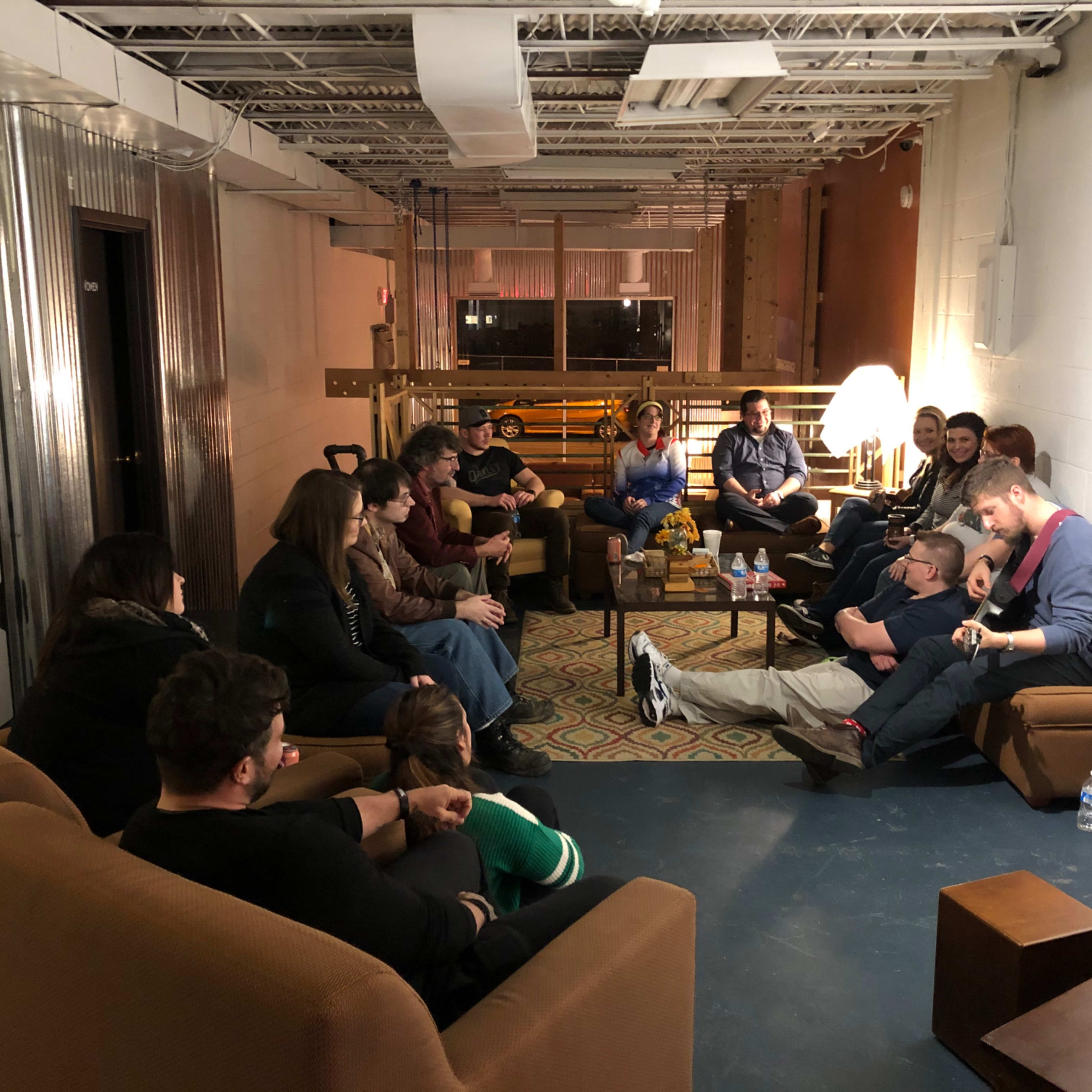 A group of people sits in a cozy lounge area, arranged in a circle around a low coffee table.