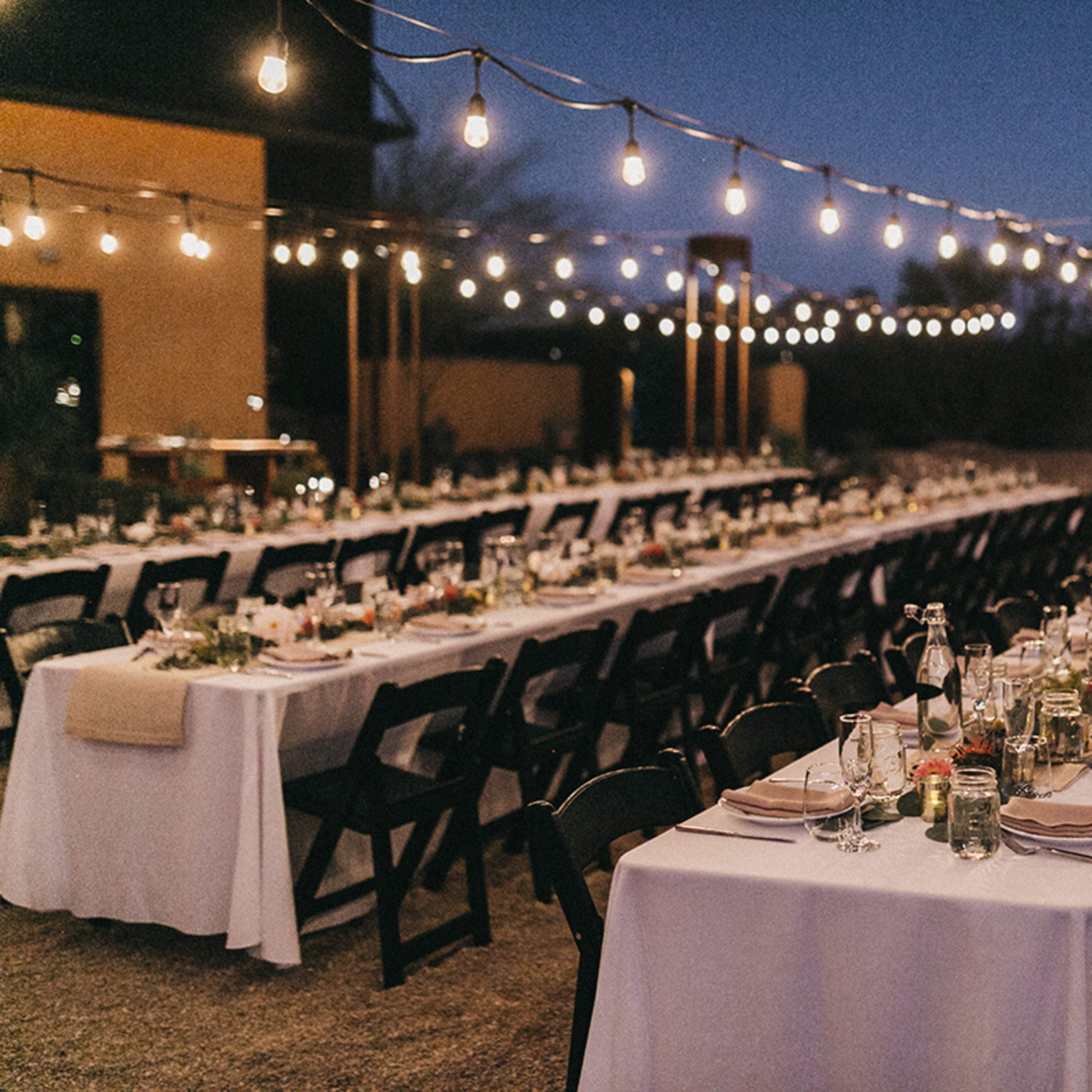A long outdoor table setup for a dinner event is illuminated by string lights against a twilight sky.
