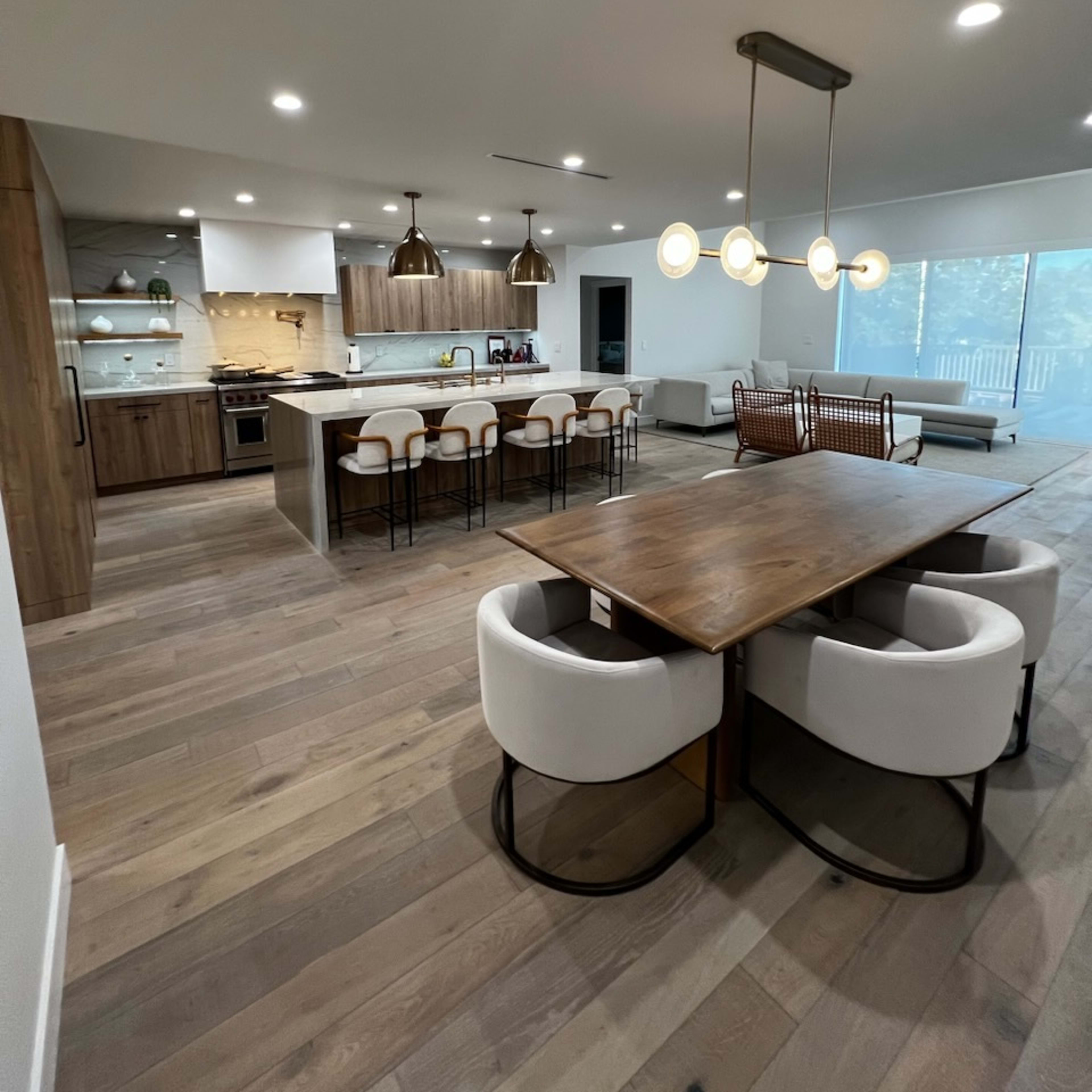 A modern kitchen and dining area with wooden flooring, featuring a large dining table, white upholstered chairs, a kitchen island with bar stools, and stylish pendant lighting.