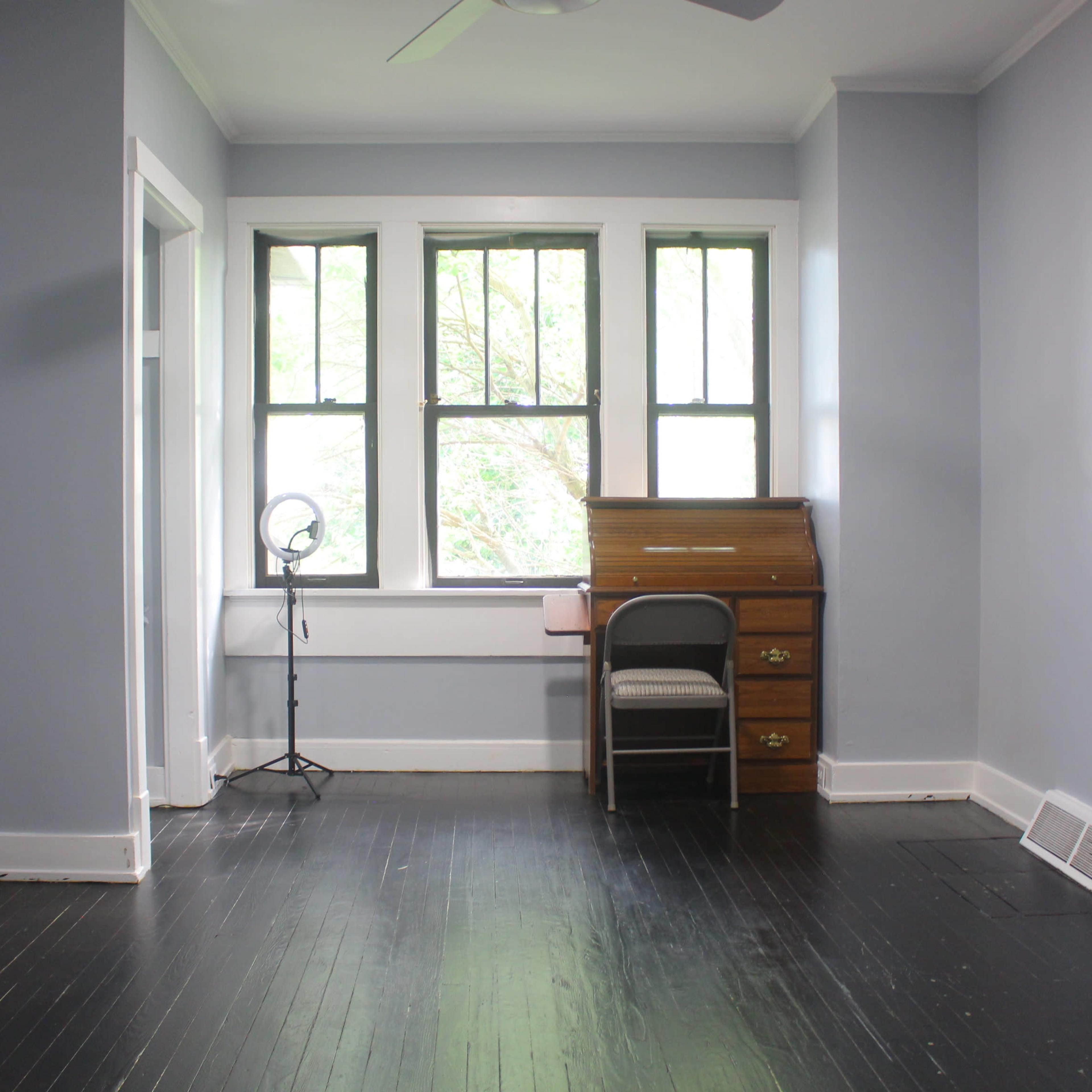 A light blue room features a wooden desk with a chair, a floor lamp, and windows overlooking greenery.