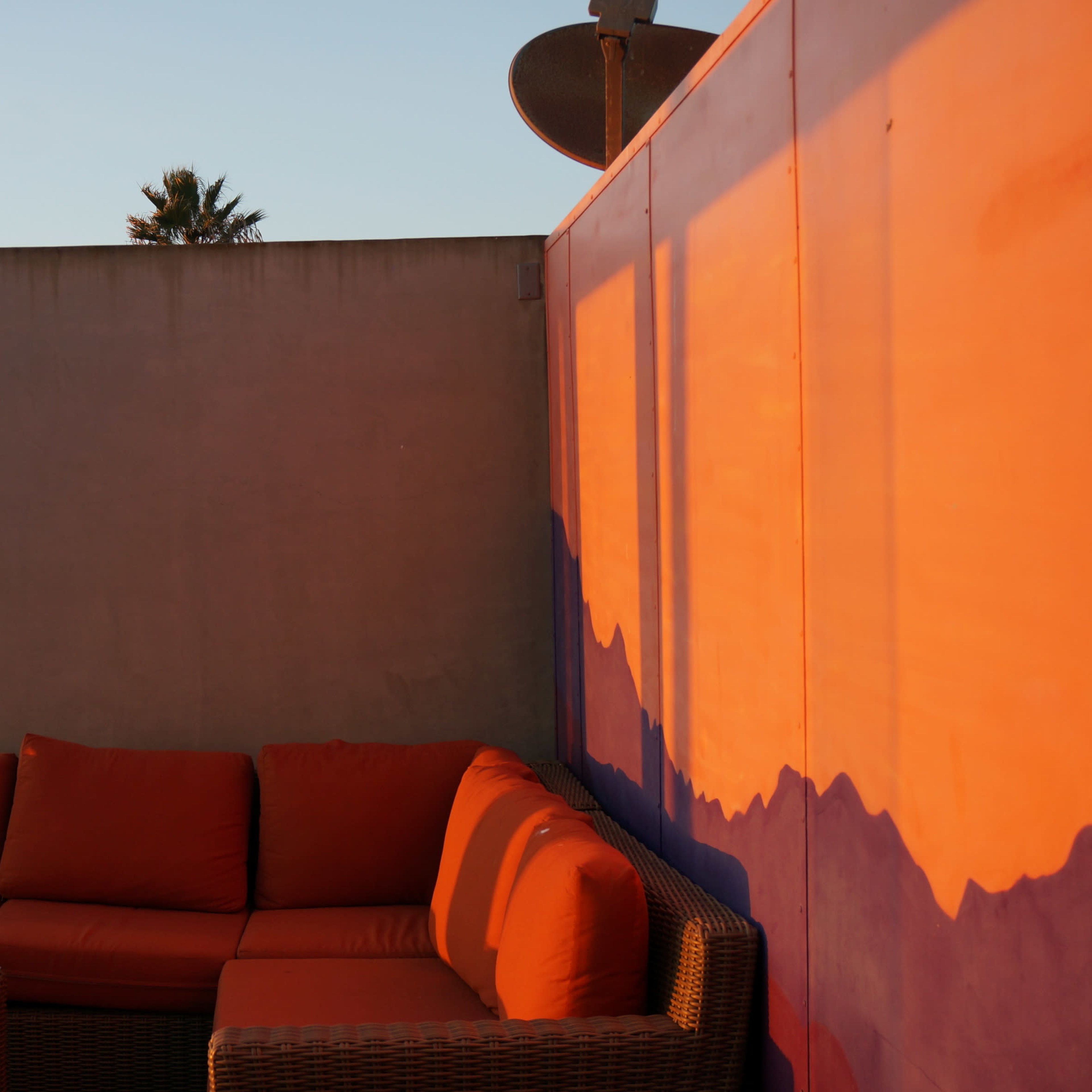 A rooftop patio features an orange sofa set against a wall with a view of palm trees and the ocean in the background.