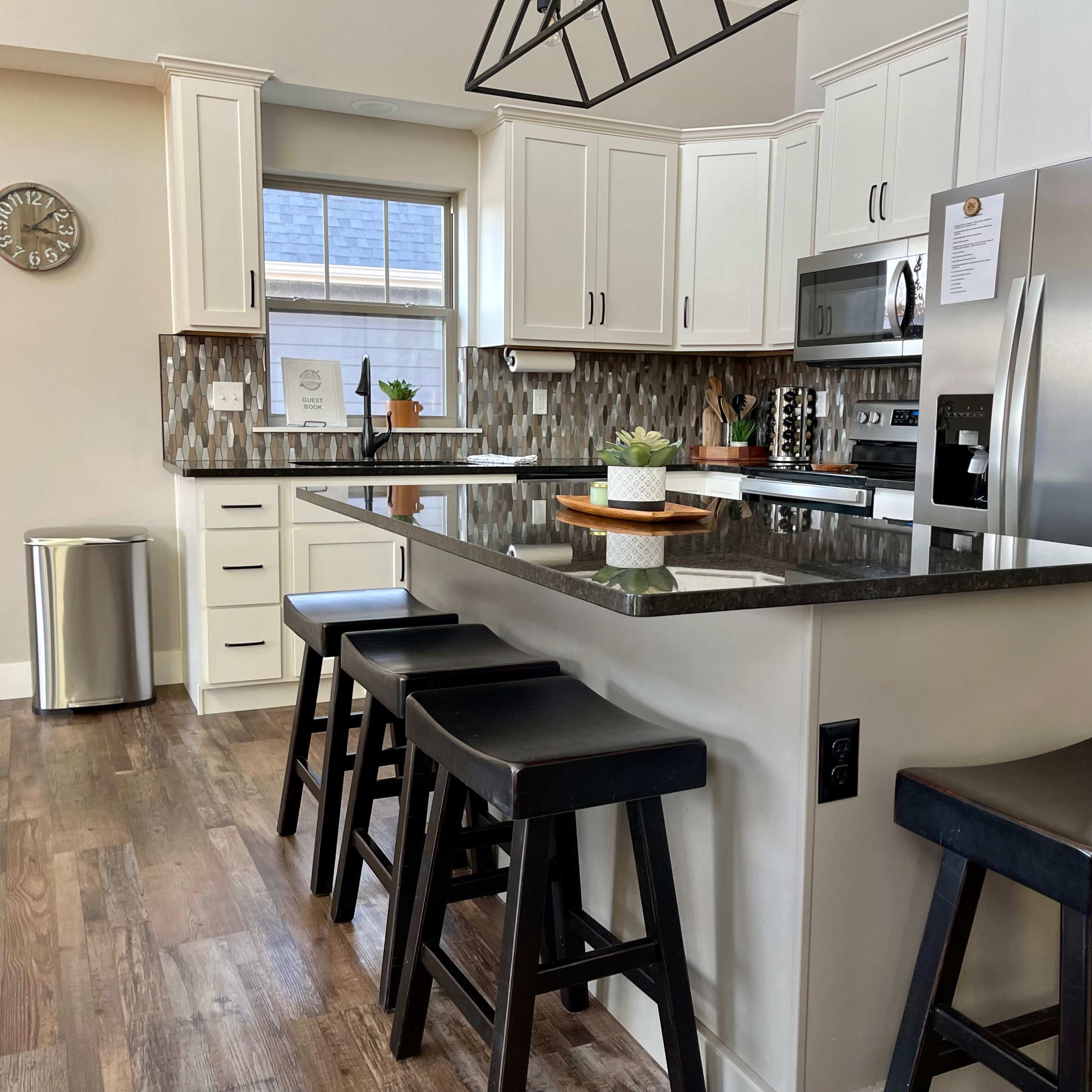 A modern kitchen with white cabinetry, a large island with black stools, stainless steel appliances, and a decorative backsplash.