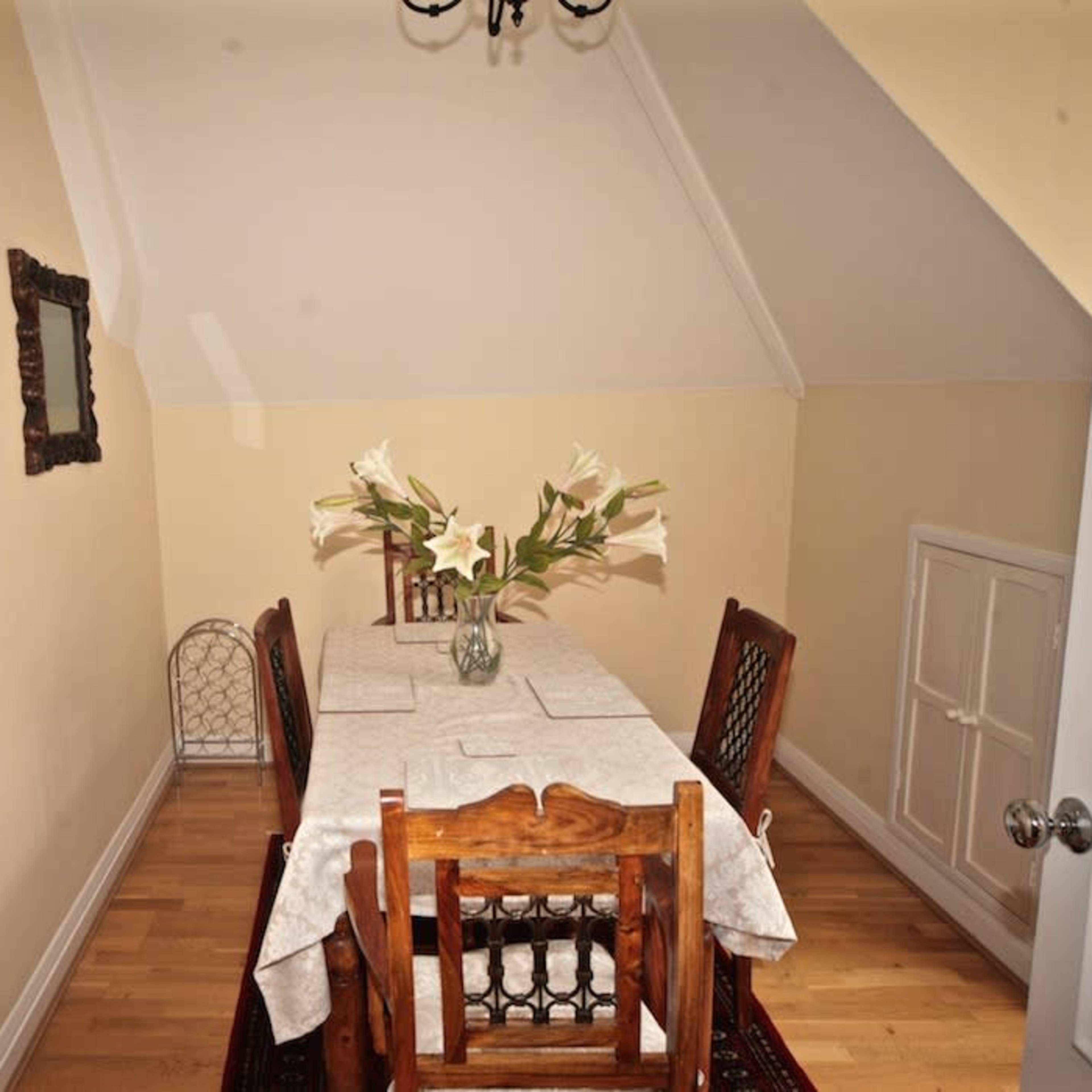 A small dining area features a wooden table surrounded by four chairs, with a vase of lilies on the table, and walls adorned with framed pictures.