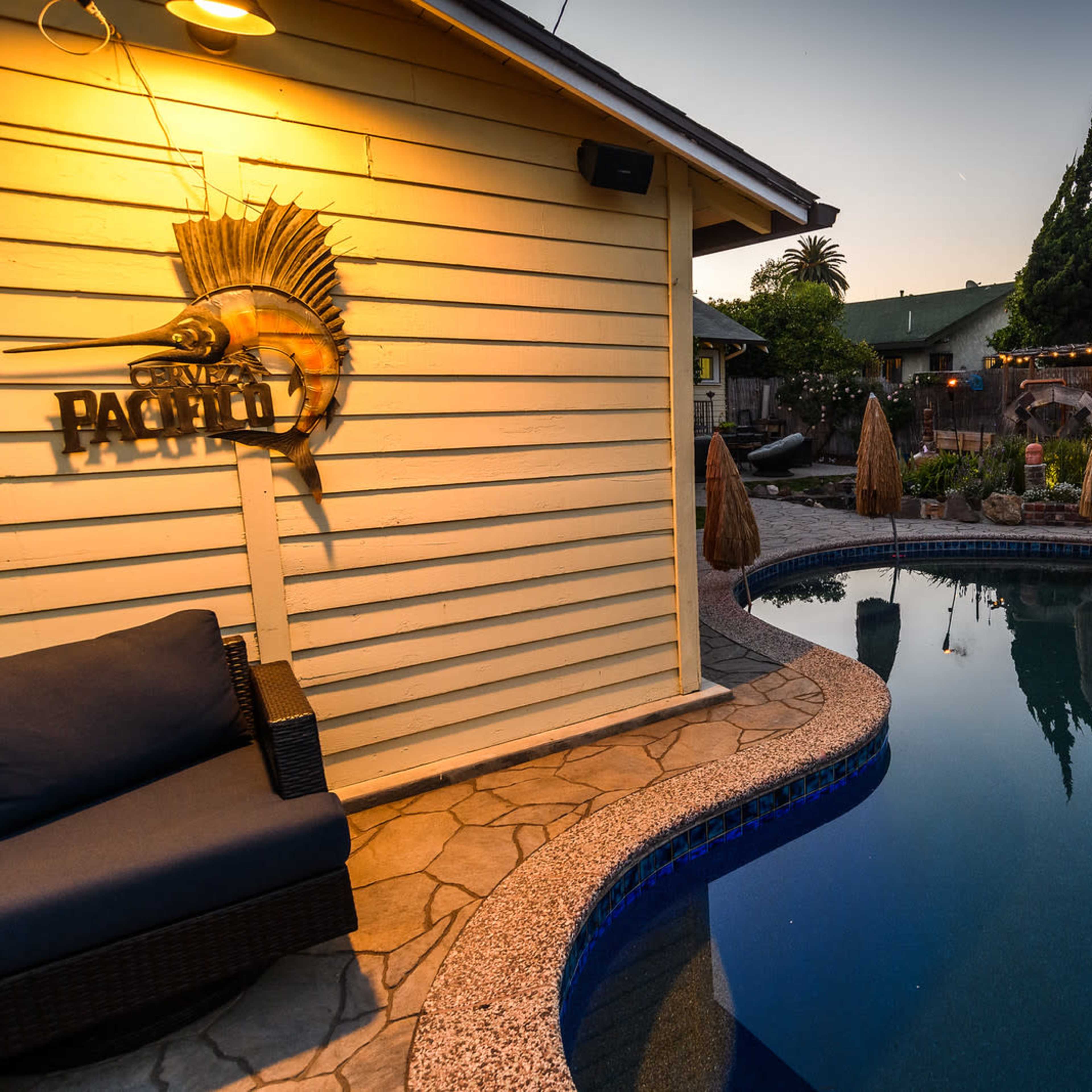 A backyard pool area with a blue swimming pool, lounge chairs, and decorative lighting in the early evening.