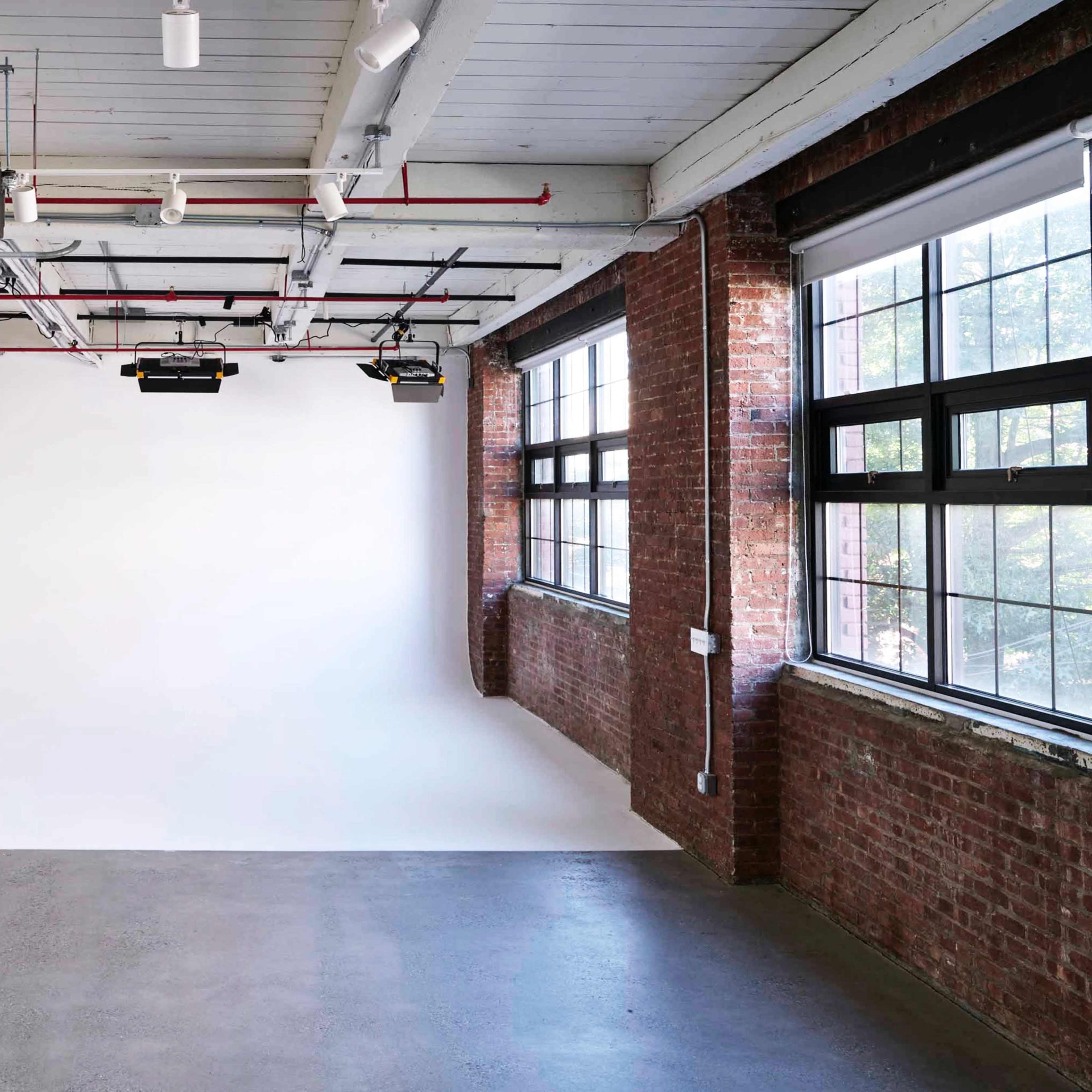 A spacious studio with a white backdrop and large windows, featuring exposed brick walls and industrial lighting fixtures.