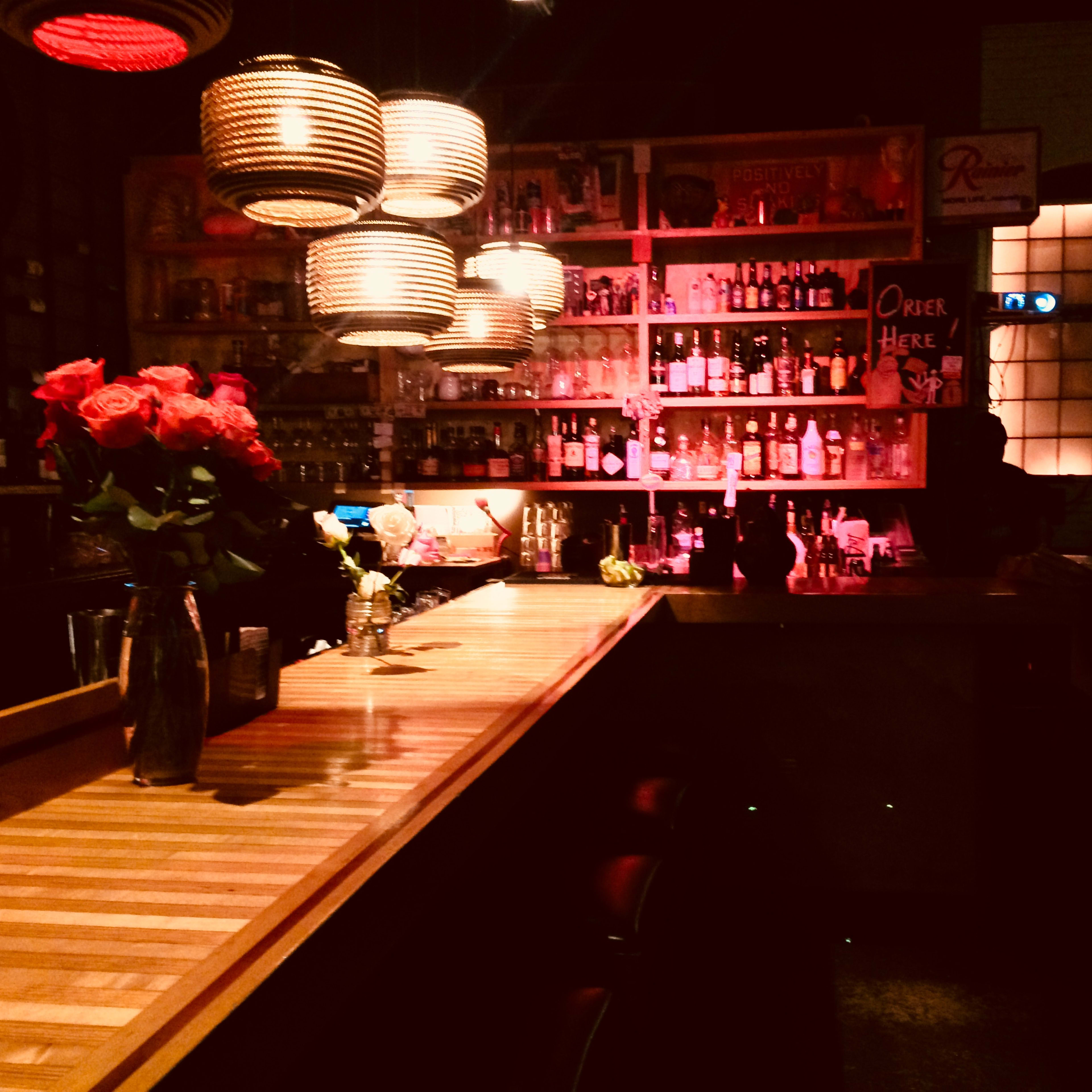 A dimly lit bar with a wooden counter, shelves lined with various bottles, and a vase of roses on the bar.