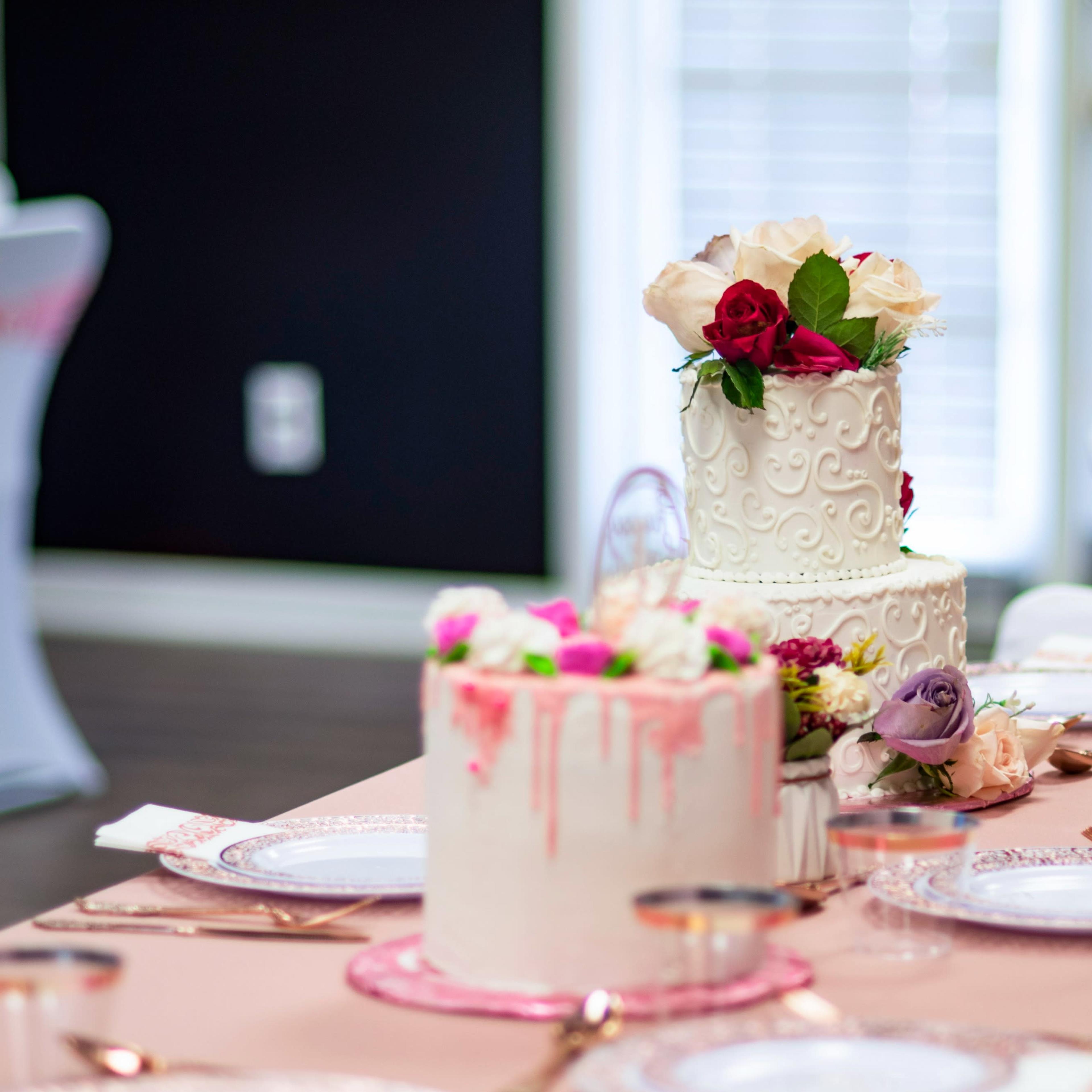 Two elegantly designed wedding cakes with decorative flowers sit on a table set with fine dishware and a pink tablecloth.