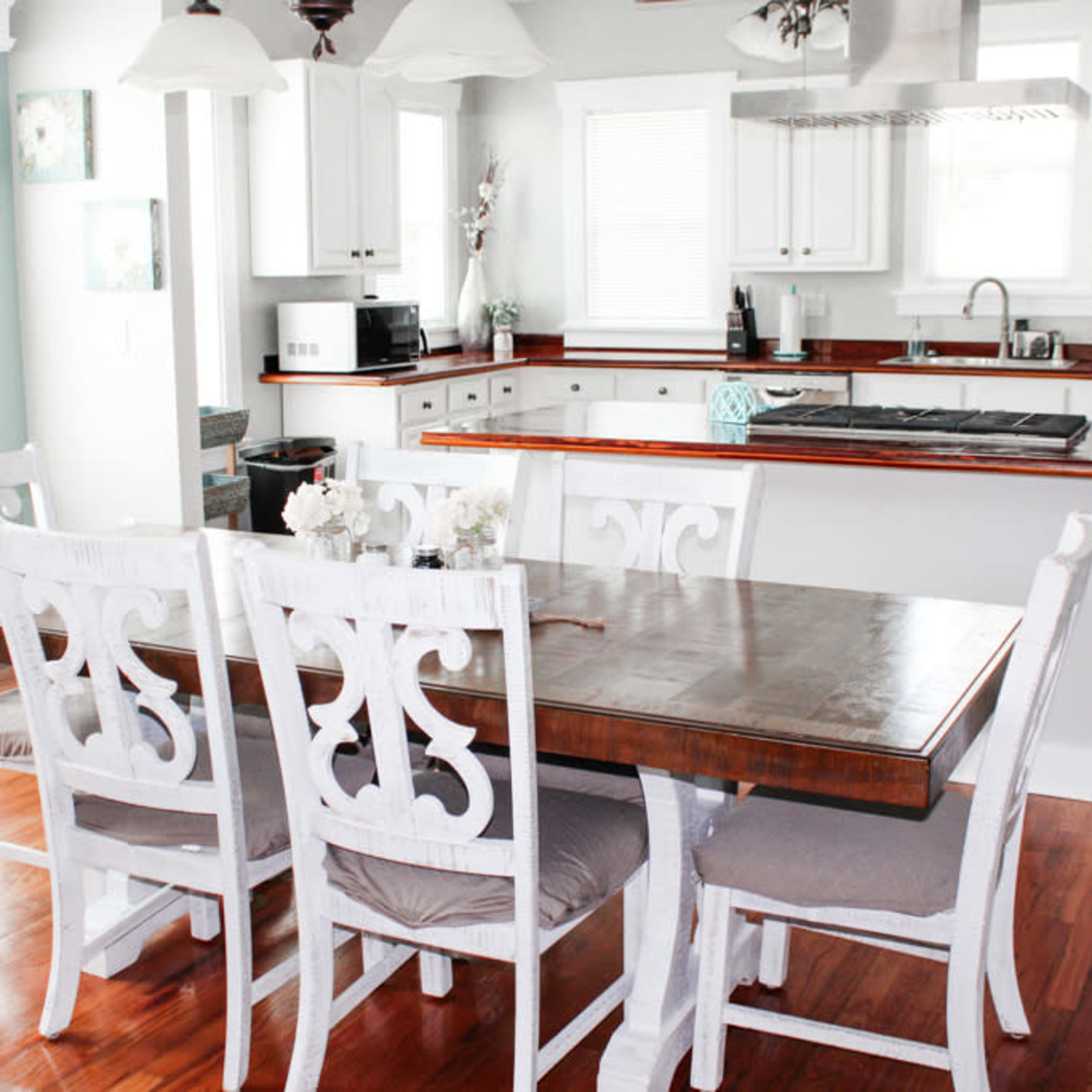 A dining table with six white chairs is set in a kitchen with wooden accents and light-colored cabinetry.
