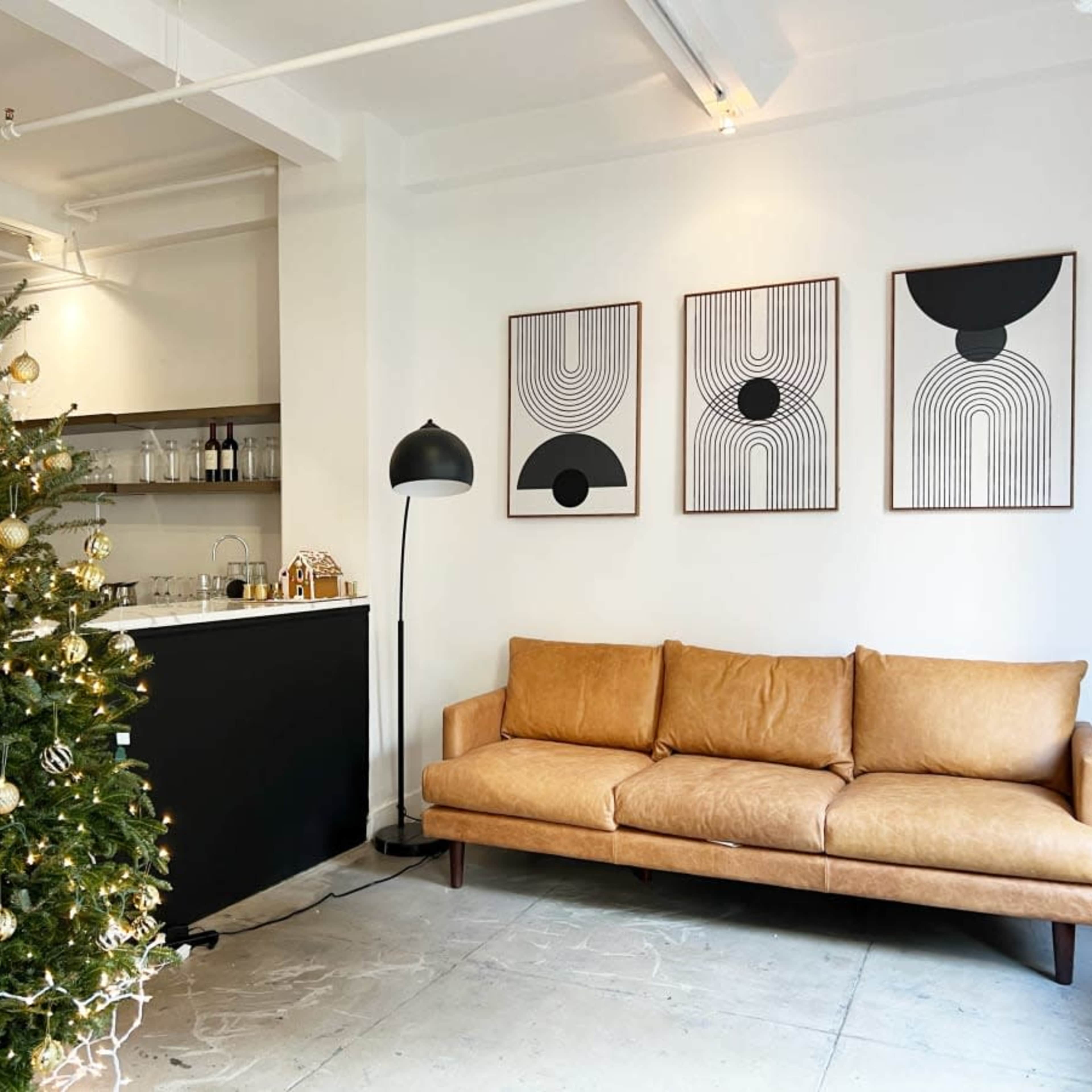 A decorated Christmas tree stands next to a brown leather sofa and three abstract framed artworks on a white wall in a modern living space.