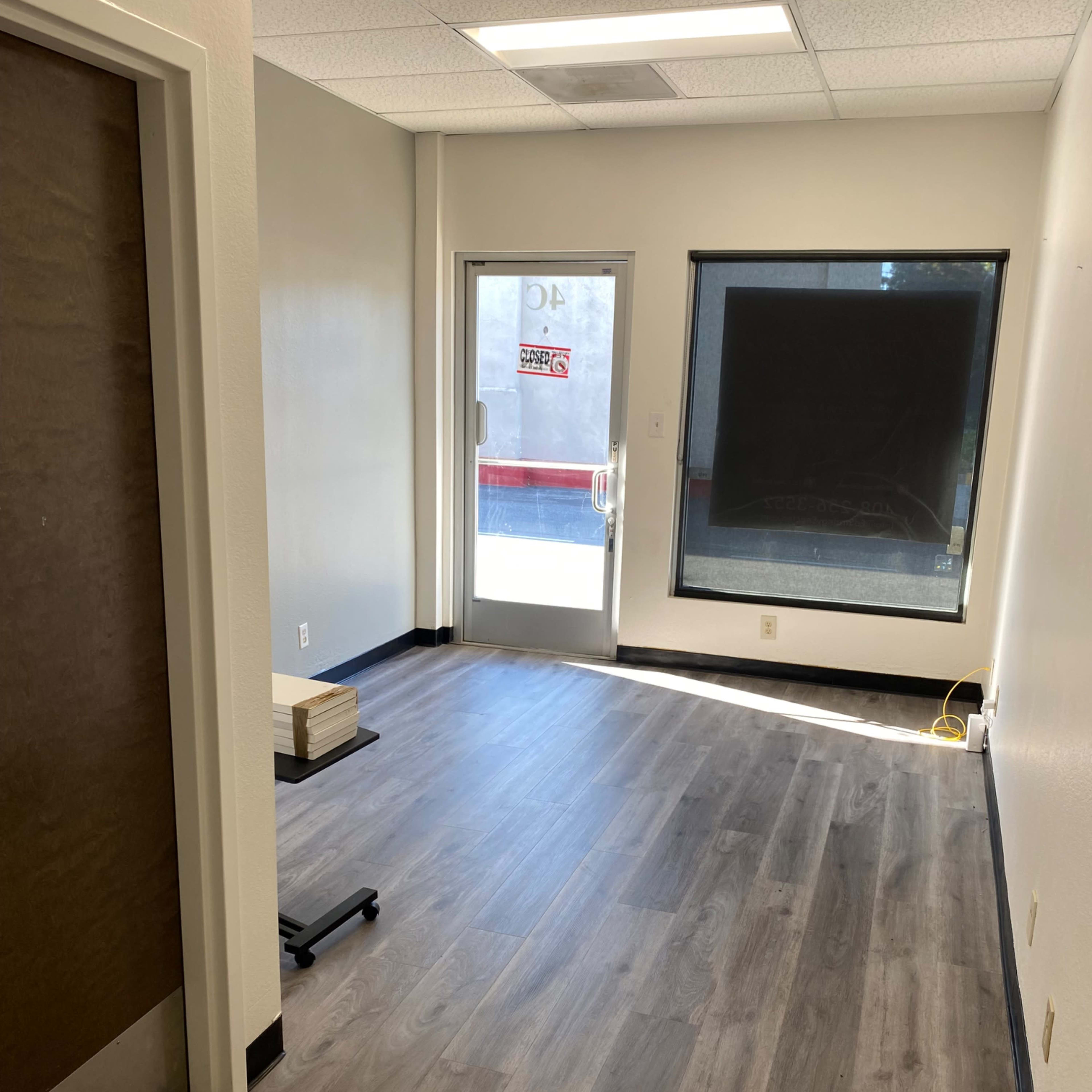An empty room with light-colored walls, a door leading outside, and large windows, featuring gray laminate flooring.