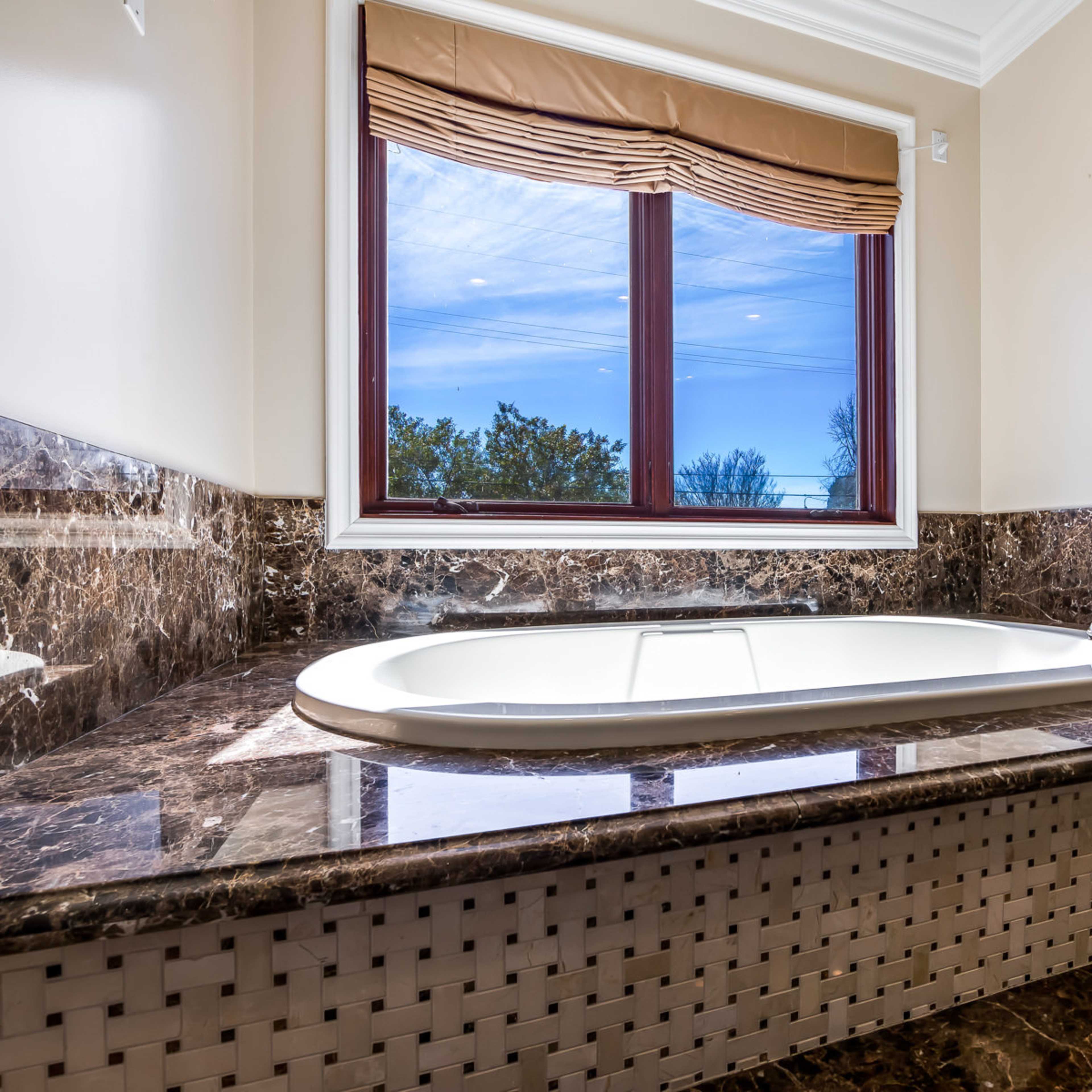 A modern bathroom featuring a large white bathtub set against a marble countertop with a window overlooking a tree-lined view.