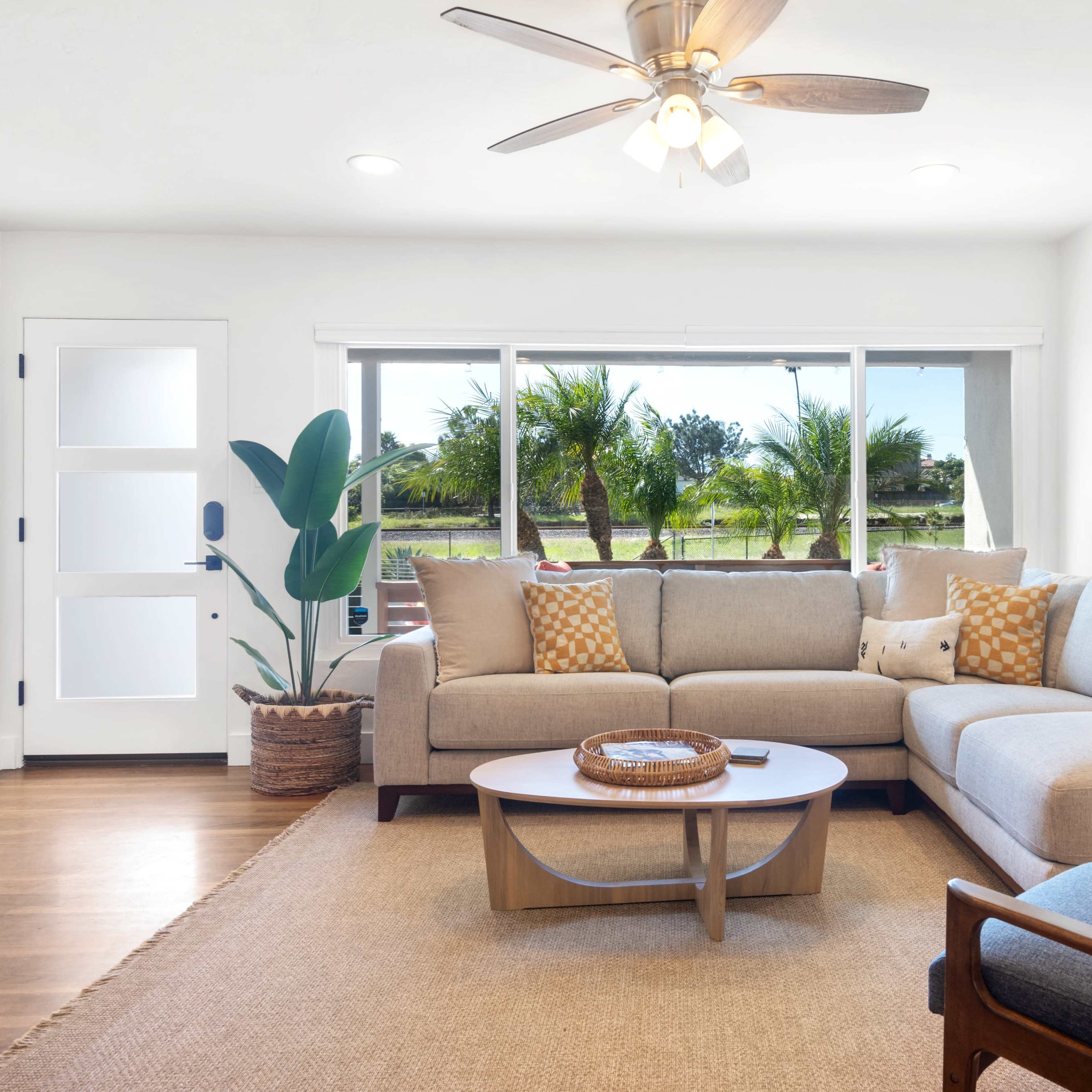 A bright living room features a sectional sofa, a round coffee table, and large windows overlooking a landscaped outdoor area with palm trees.