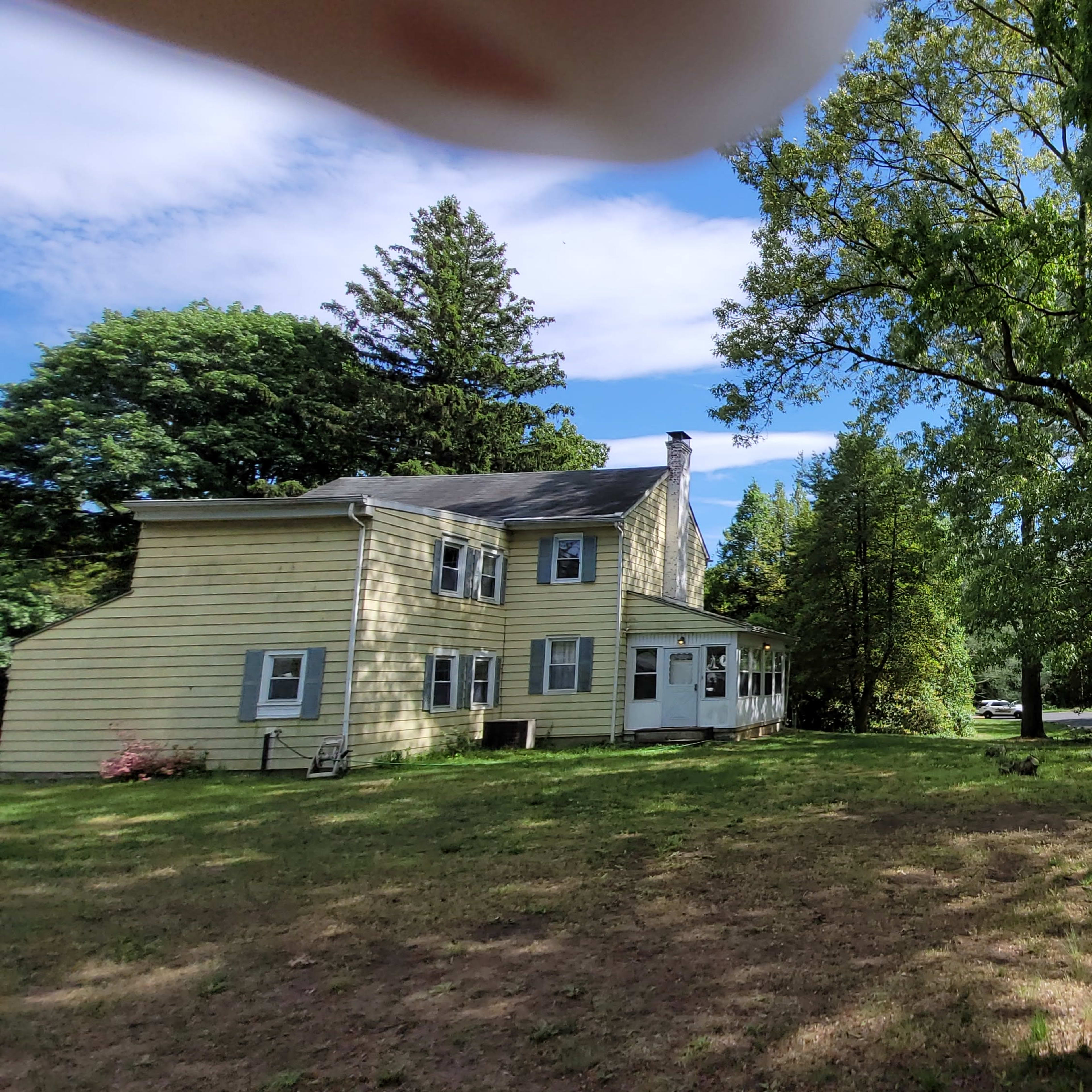 A yellow two-story house is situated on a grassy area surrounded by tall trees under a partly cloudy sky.