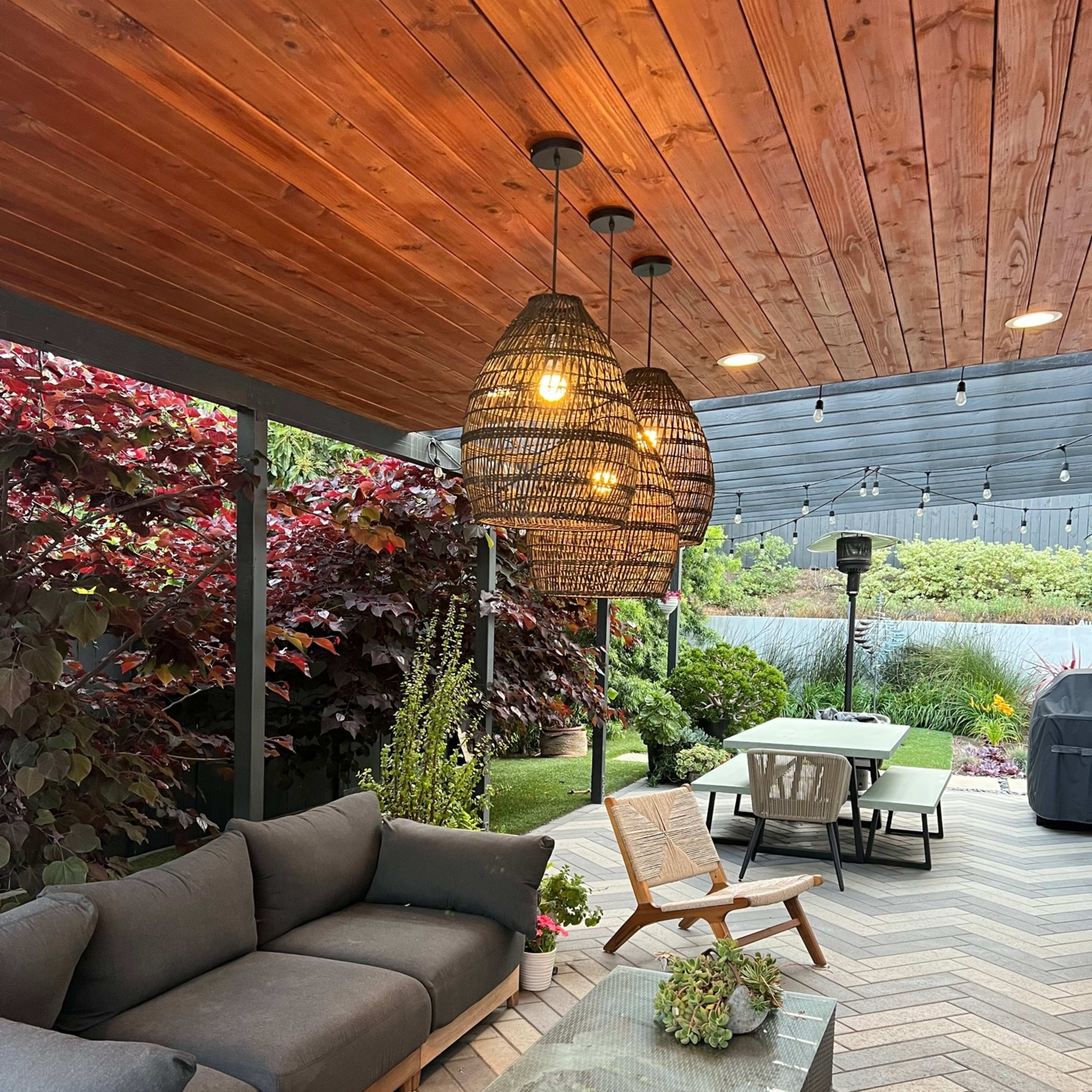 A cozy outdoor patio area featuring a sectional sofa, a glass coffee table, and three pendant lights hanging from a wooden ceiling, surrounded by lush greenery and a dining table in the background.