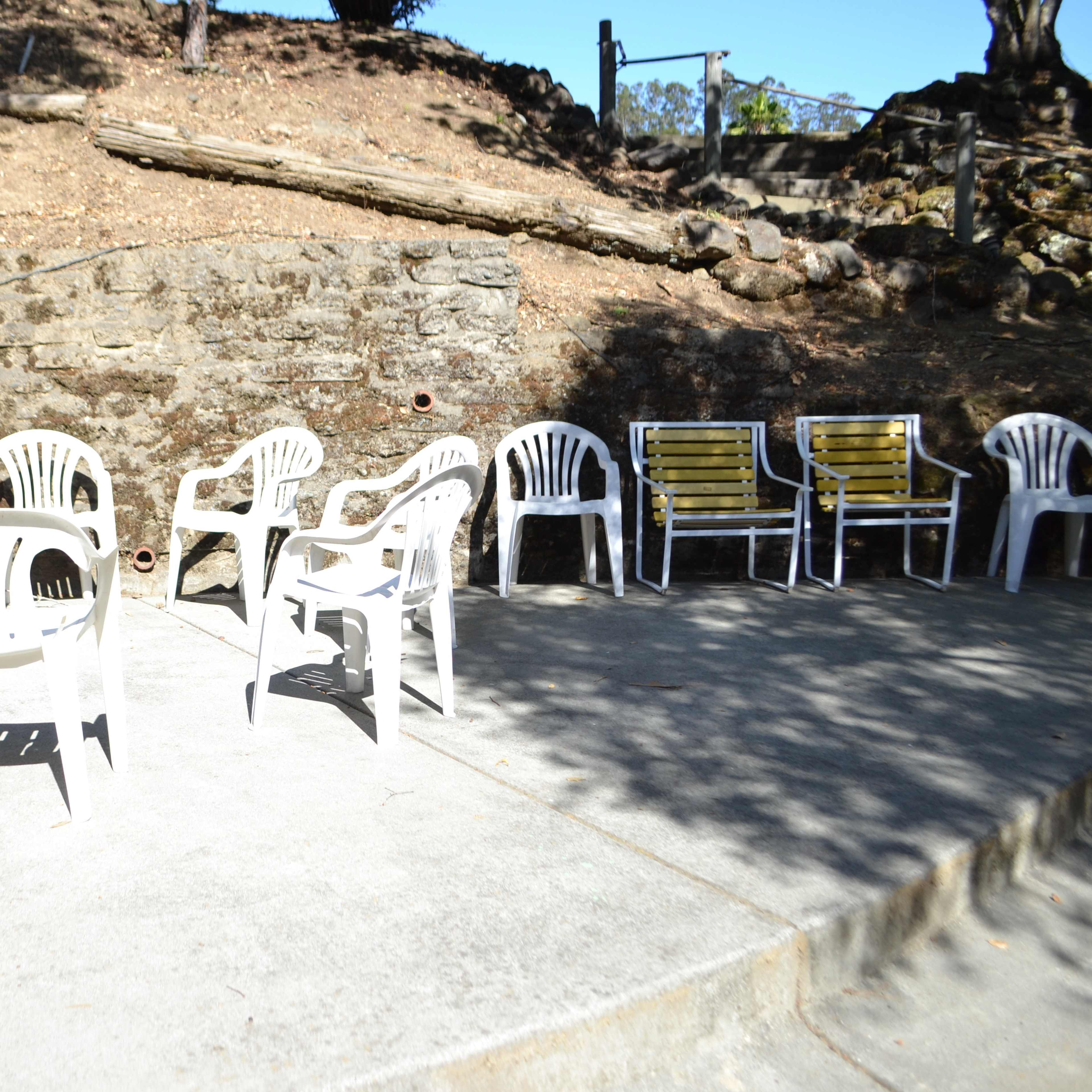A circular arrangement of white plastic chairs and tables is set on a concrete surface near a stone wall, with additional chairs positioned against the wall.