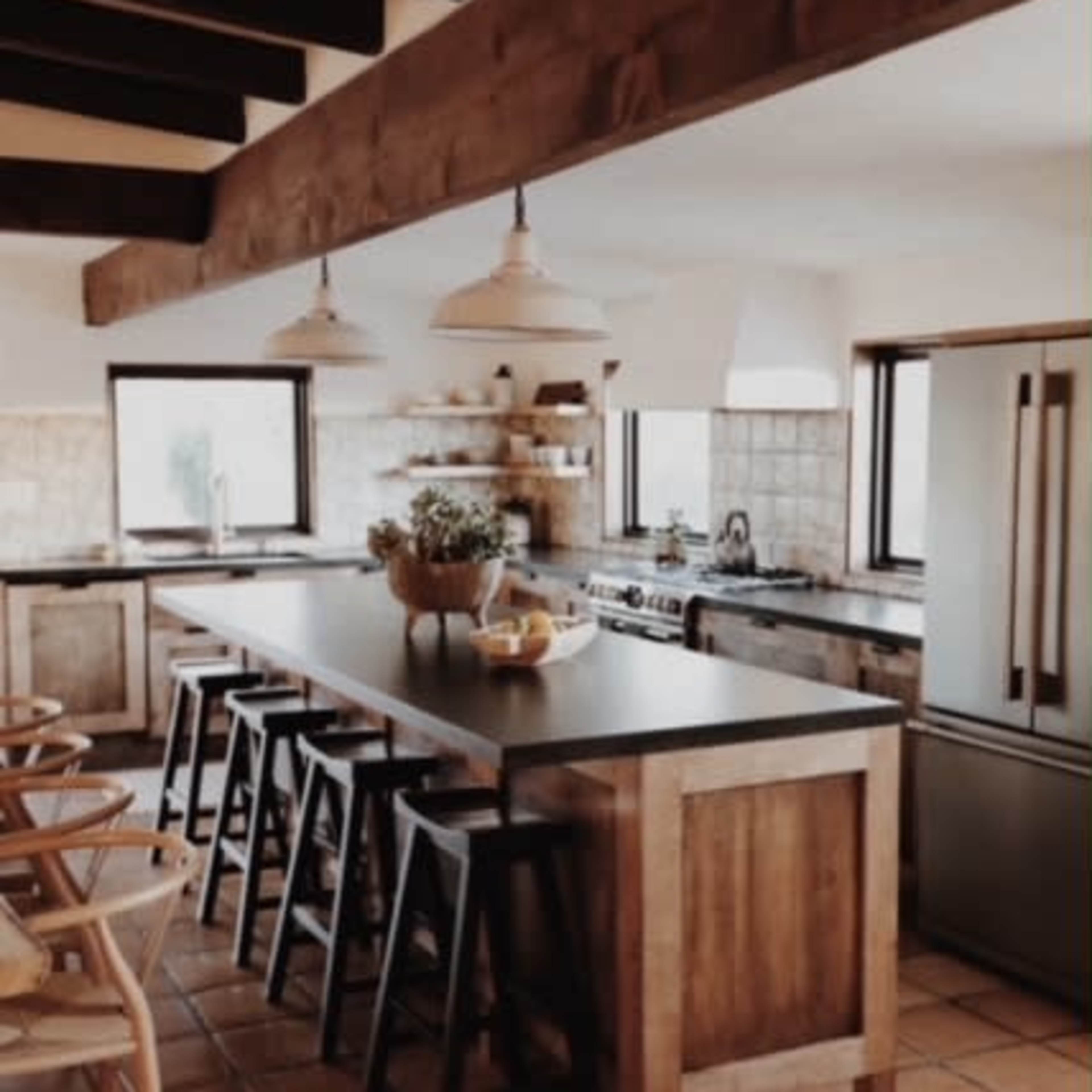 A modern kitchen features a long wooden island with black stools, exposed beams above, and large windows letting in natural light.