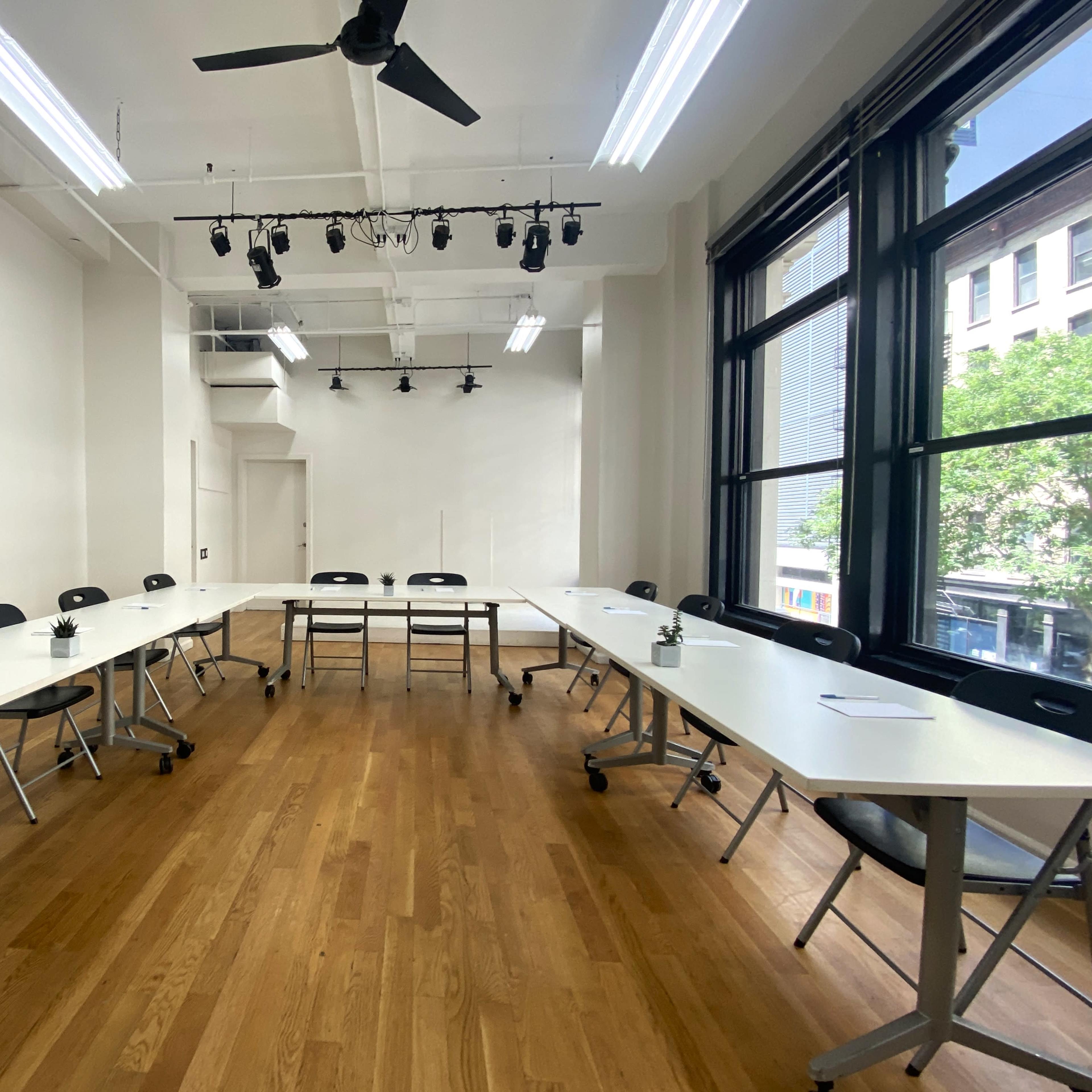 A modern conference room features a U-shaped arrangement of white tables and black chairs, with large windows allowing natural light to enter.