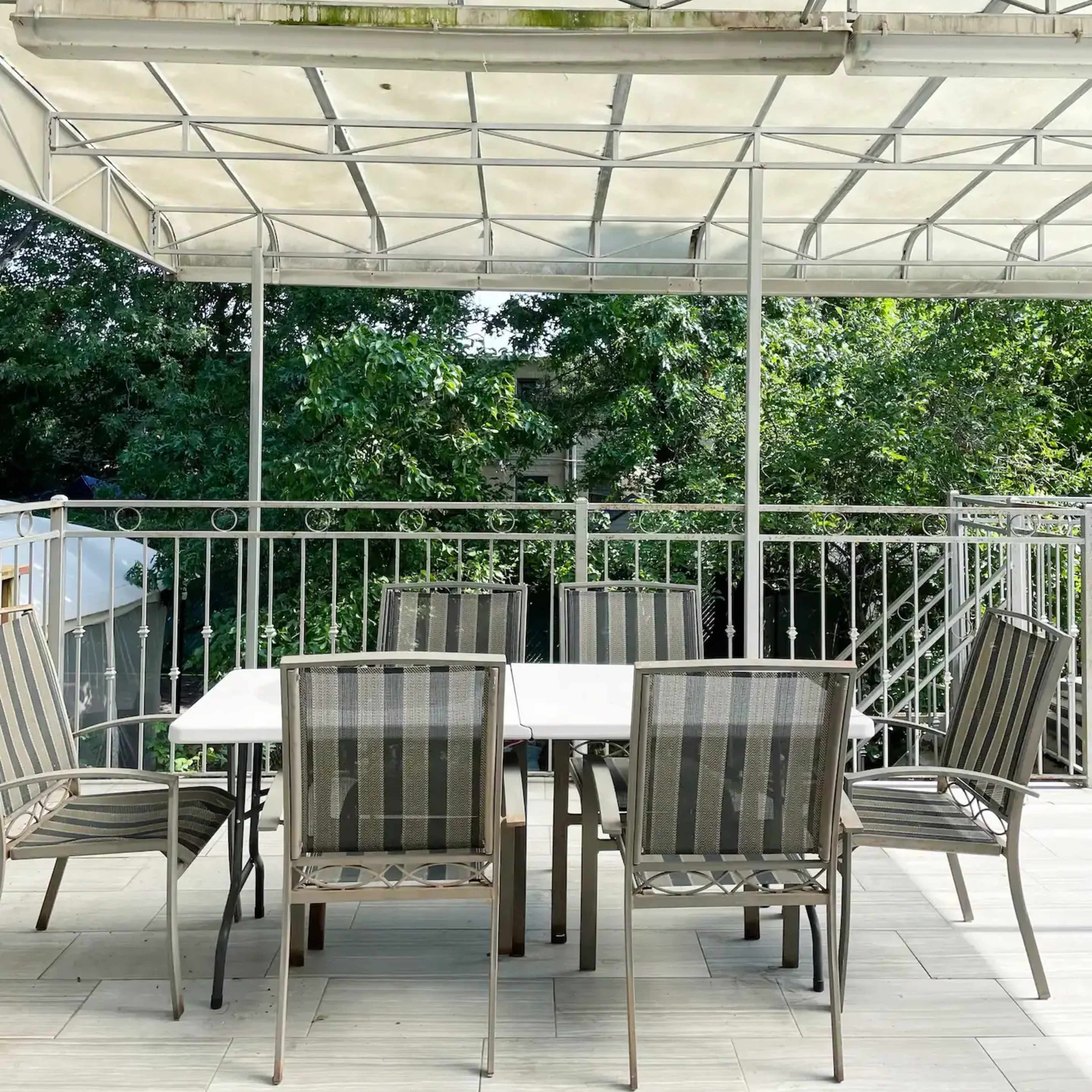 A patio with a rectangular table surrounded by six striped chairs is covered by a transparent canopy and overlooks greenery.