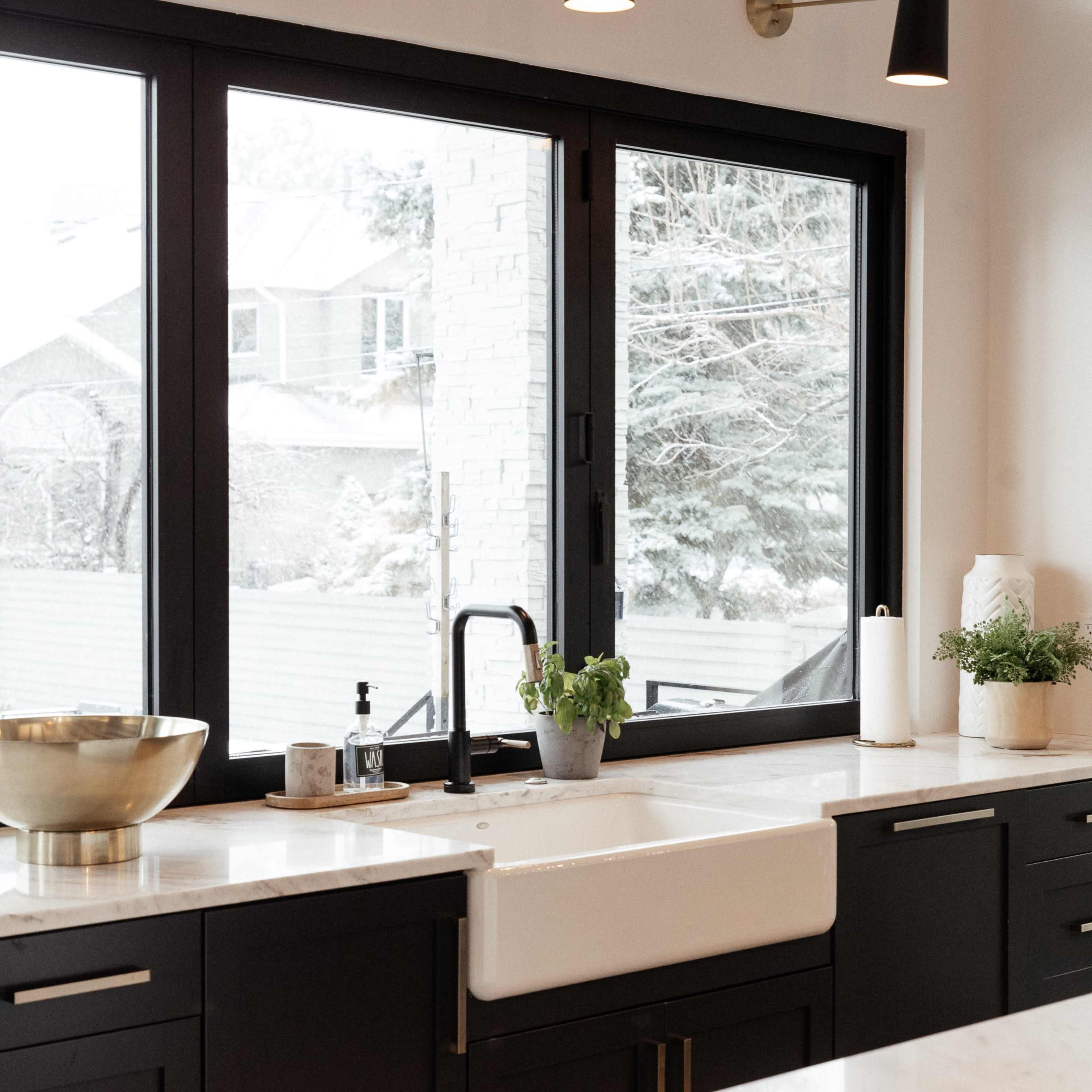 A modern kitchen featuring a farmhouse sink, black cabinetry, and large windows with a snowy landscape visible outside.