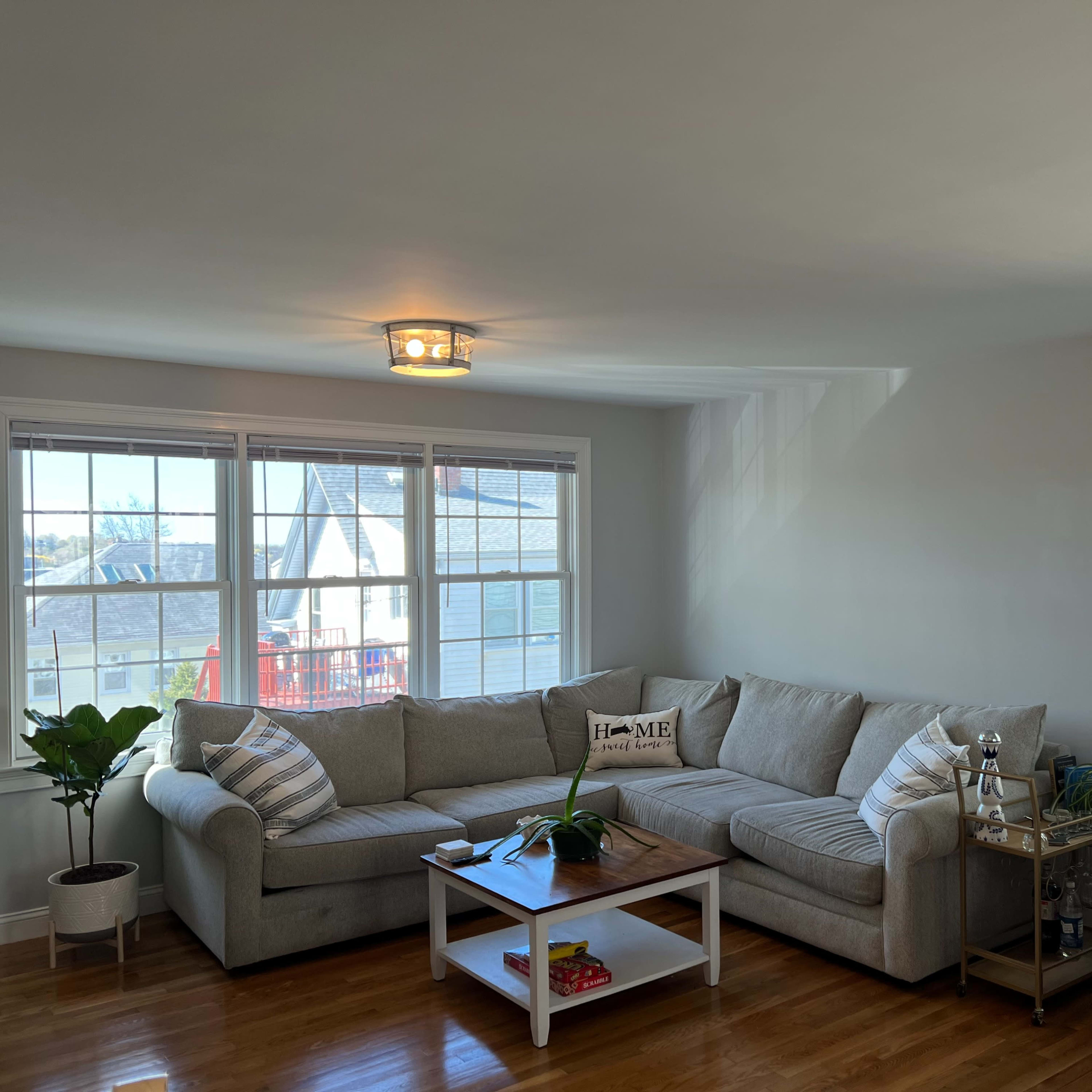 A spacious living room features a light-colored sectional sofa, a wooden coffee table, and several potted plants near large windows.