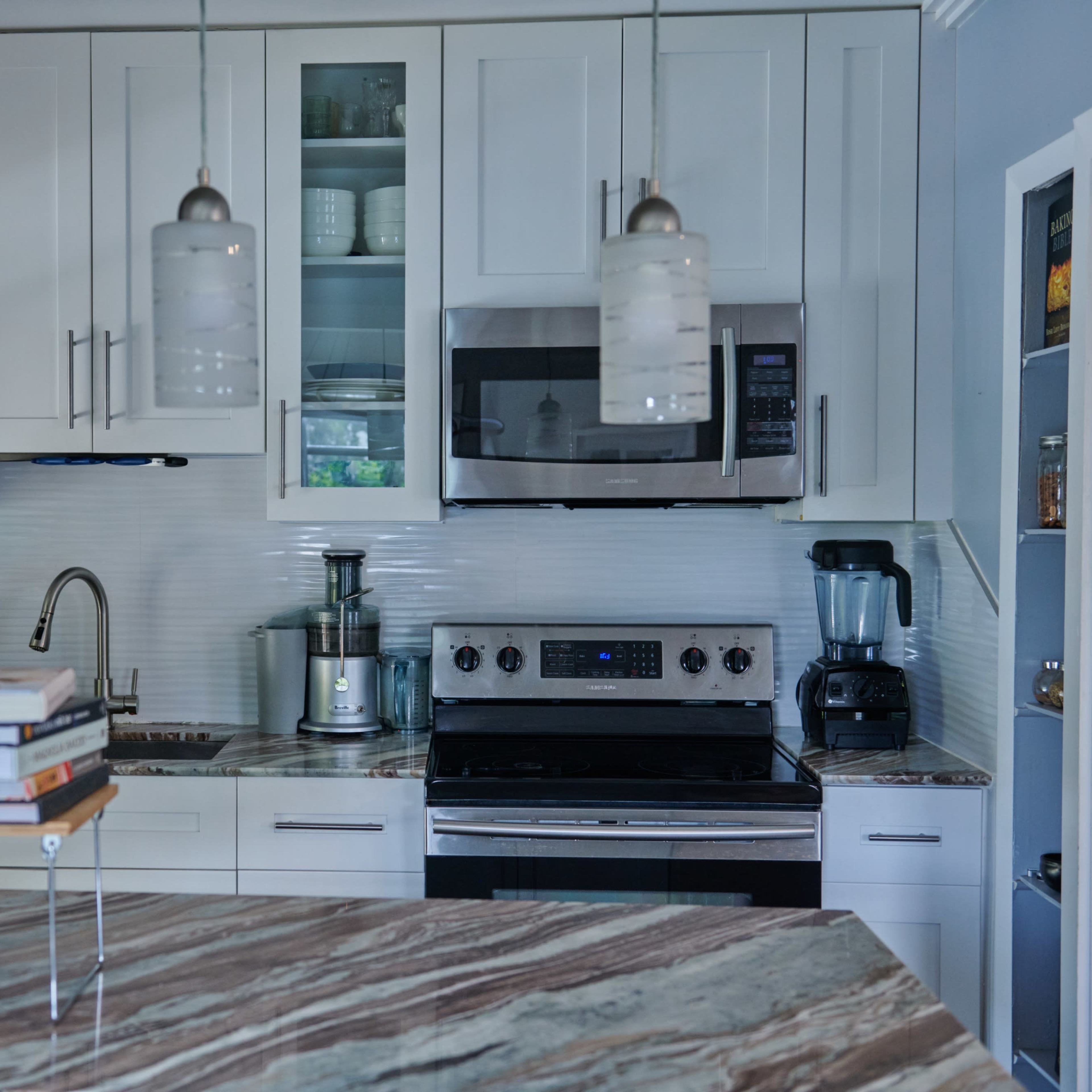 A modern kitchen features stainless steel appliances, white cabinetry, and a marble countertop with stacked cookbooks.