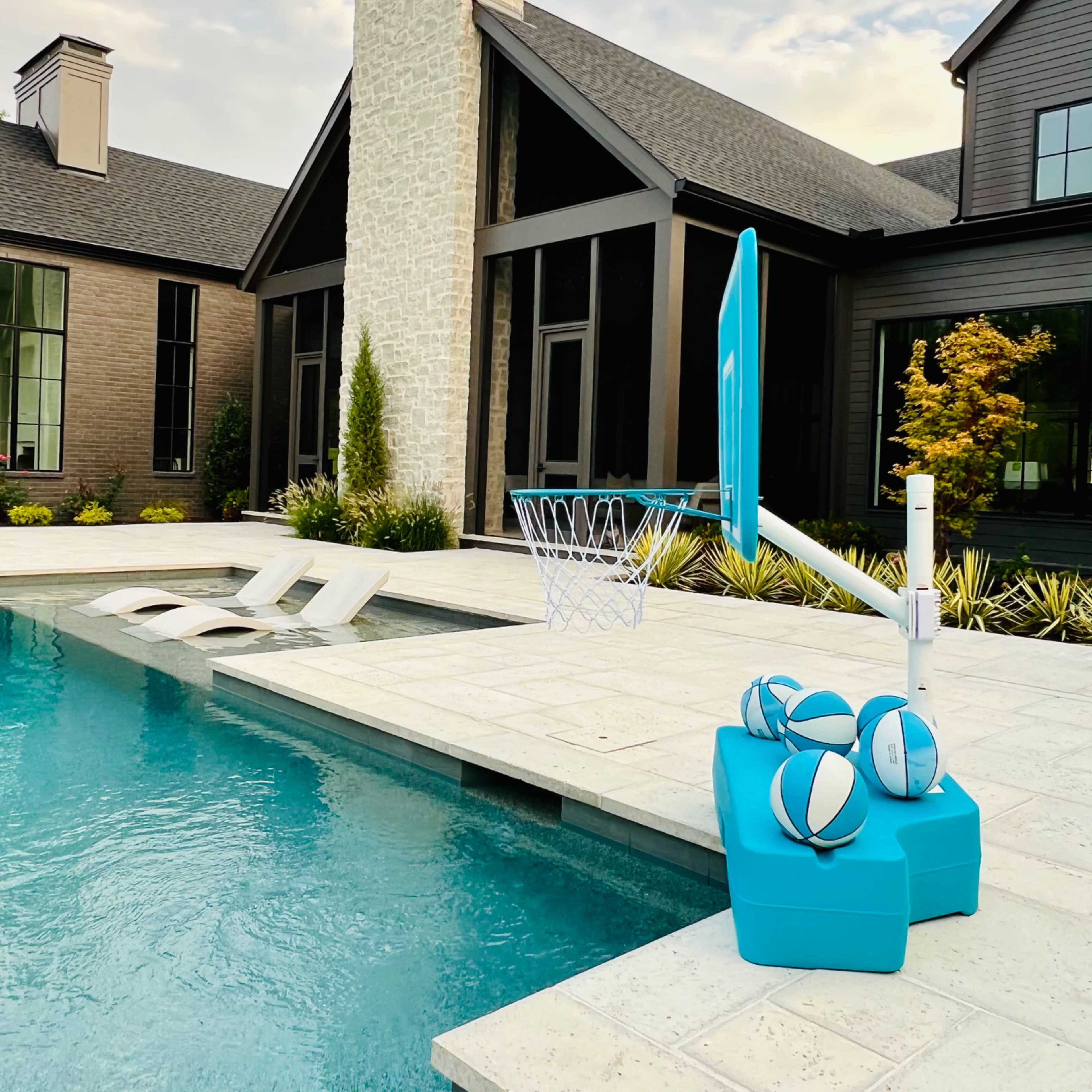 A basketball hoop stands by a swimming pool with several basketballs resting on a bright blue seating area.