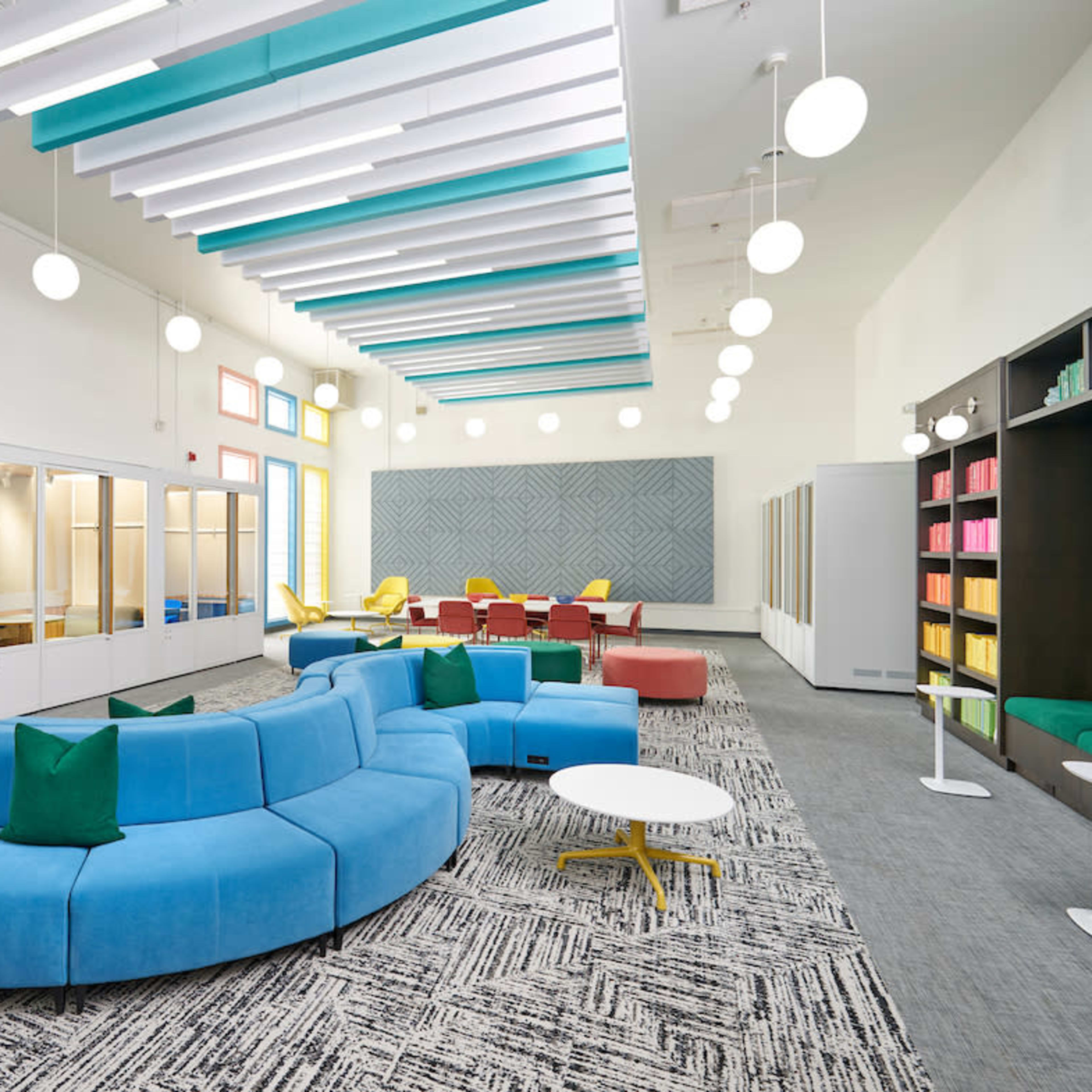 A modern, brightly lit room featuring a blue sectional sofa, various seating areas, and glass partitions for individual workspaces.