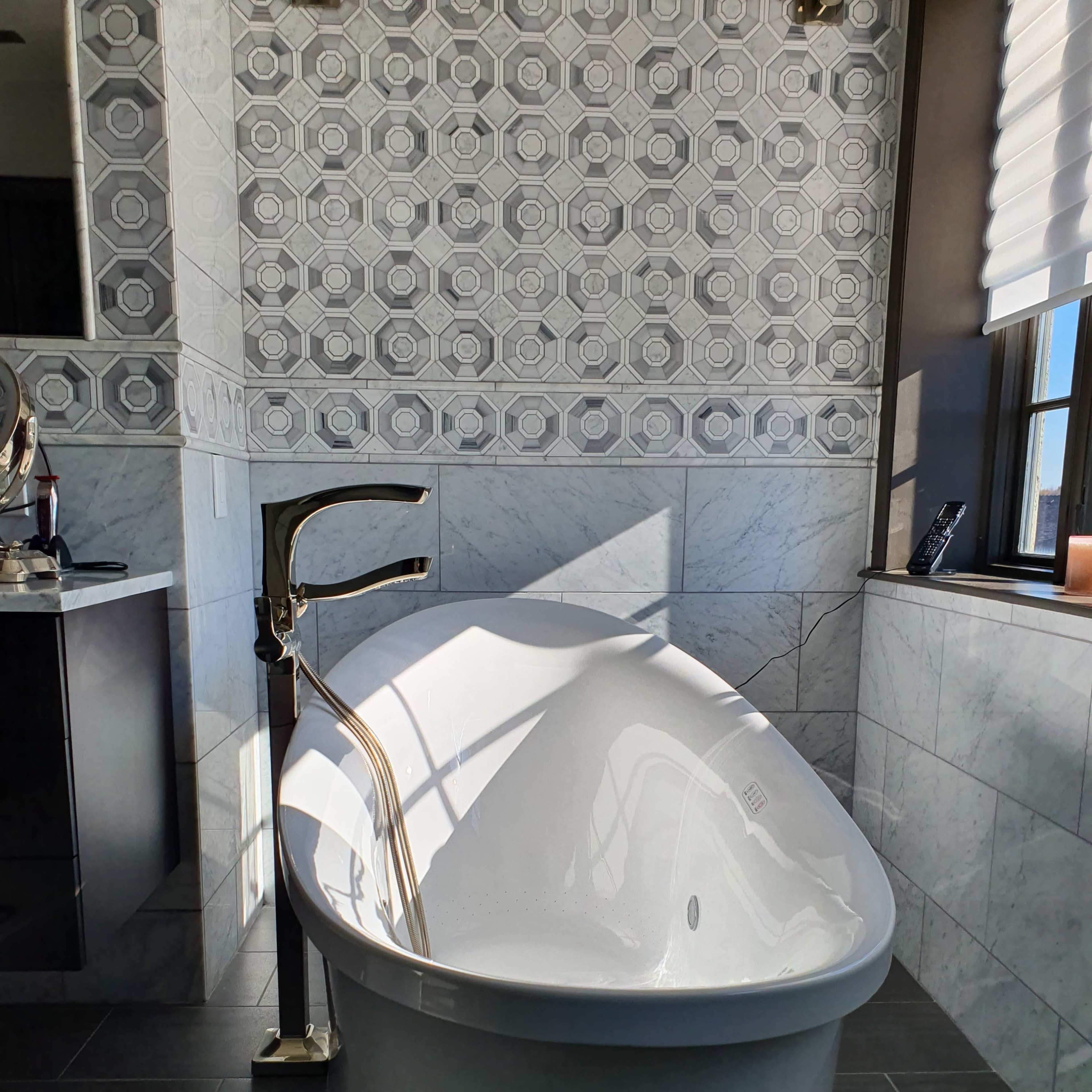 A modern bathroom features a freestanding white bathtub with a stylish faucet, set against a backdrop of geometric tiled walls and marble accents.