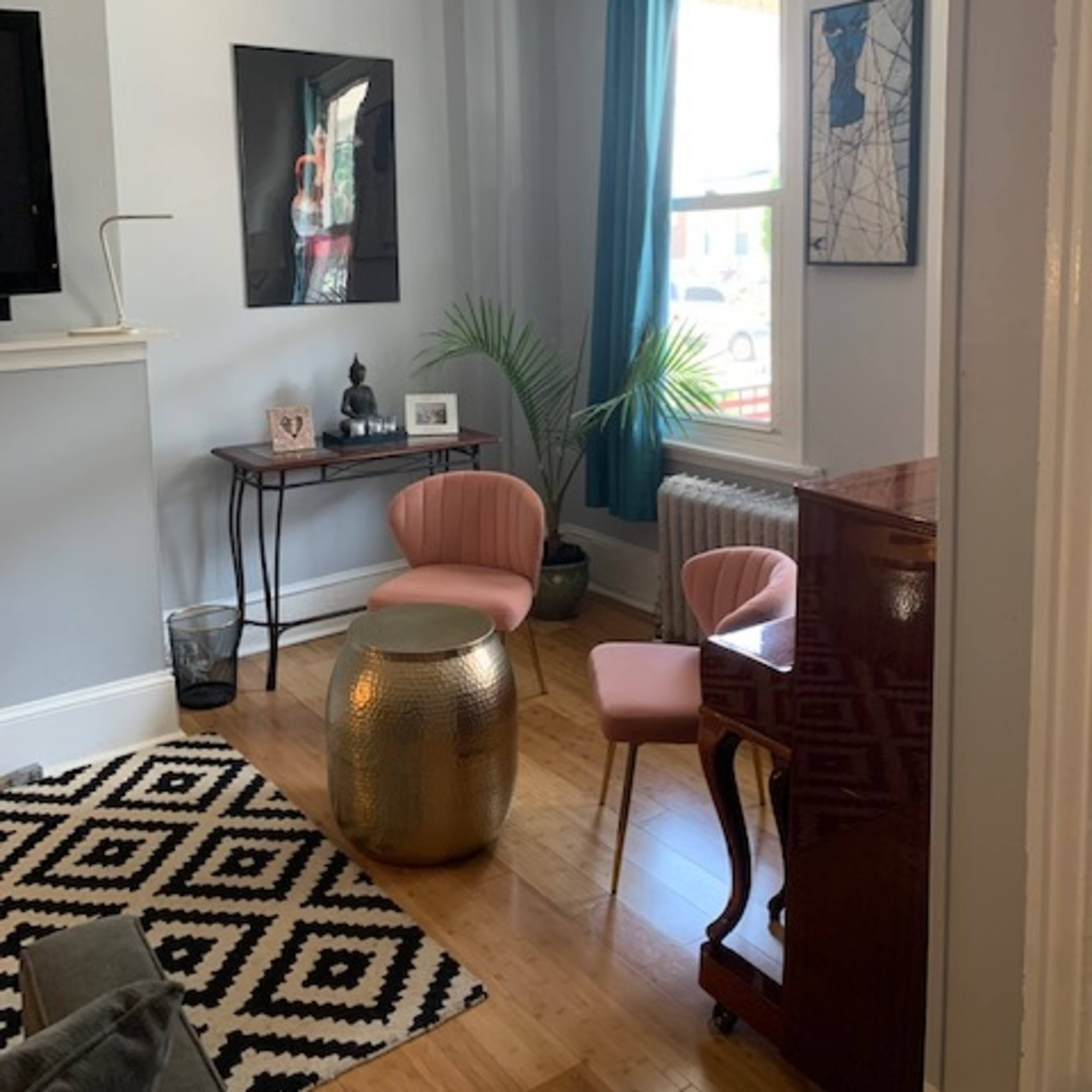 A sunny sitting area features a small table with decorative items, two pink chairs, a piano, and a patterned rug on wooden flooring.