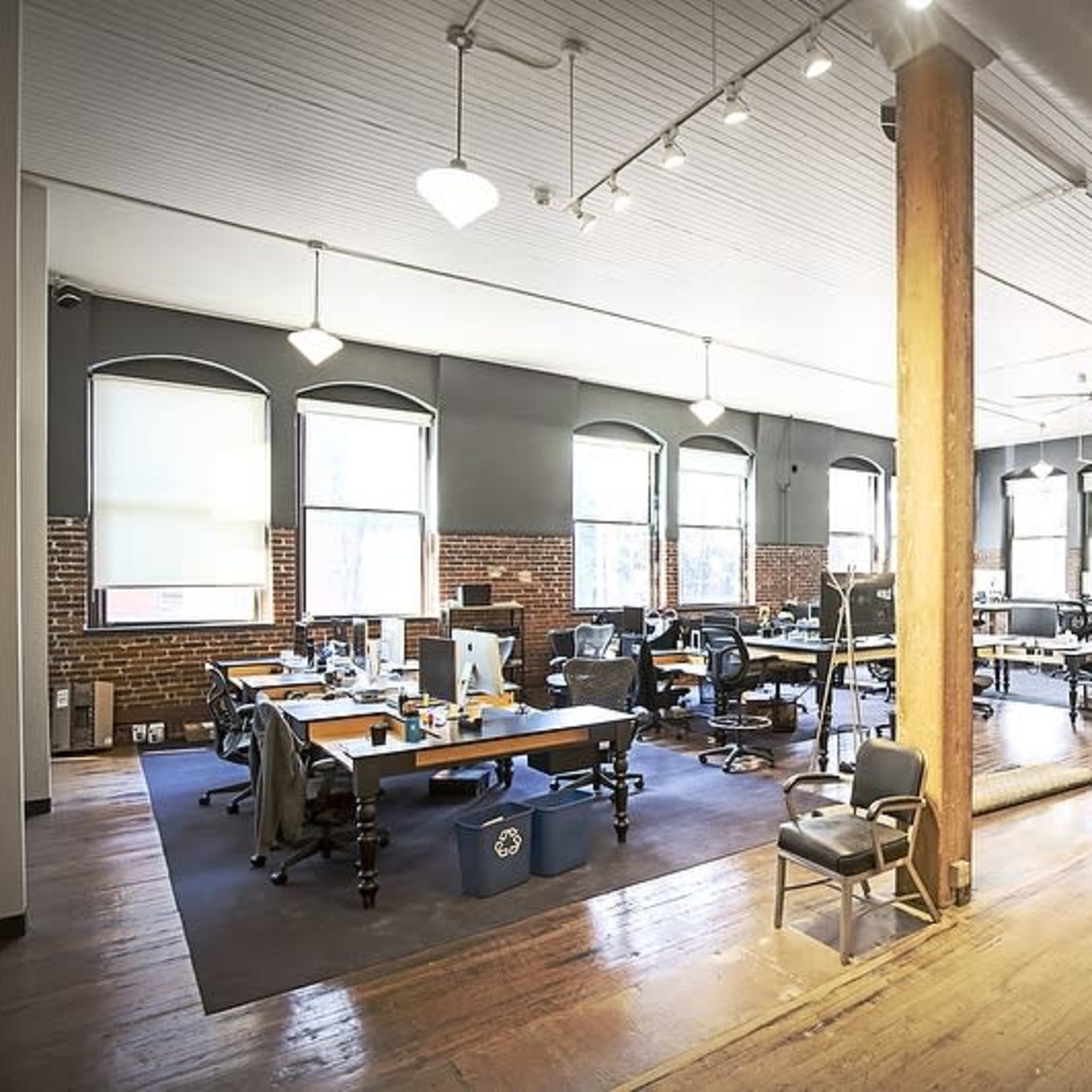A spacious office with wooden floors, large windows, and rows of desks equipped with computers.
