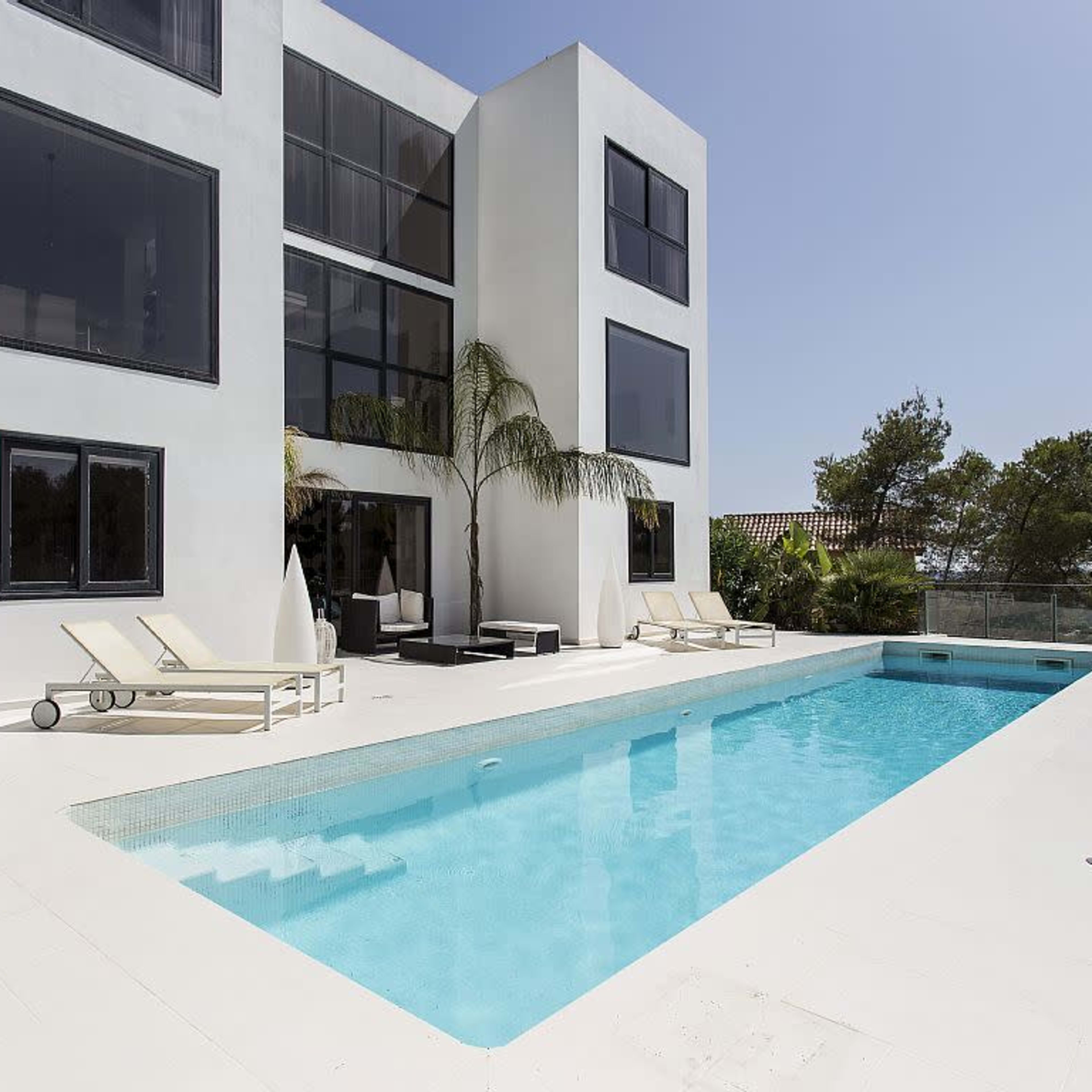 A modern white villa with large windows overlooks a rectangular swimming pool surrounded by lounge chairs and tropical plants.