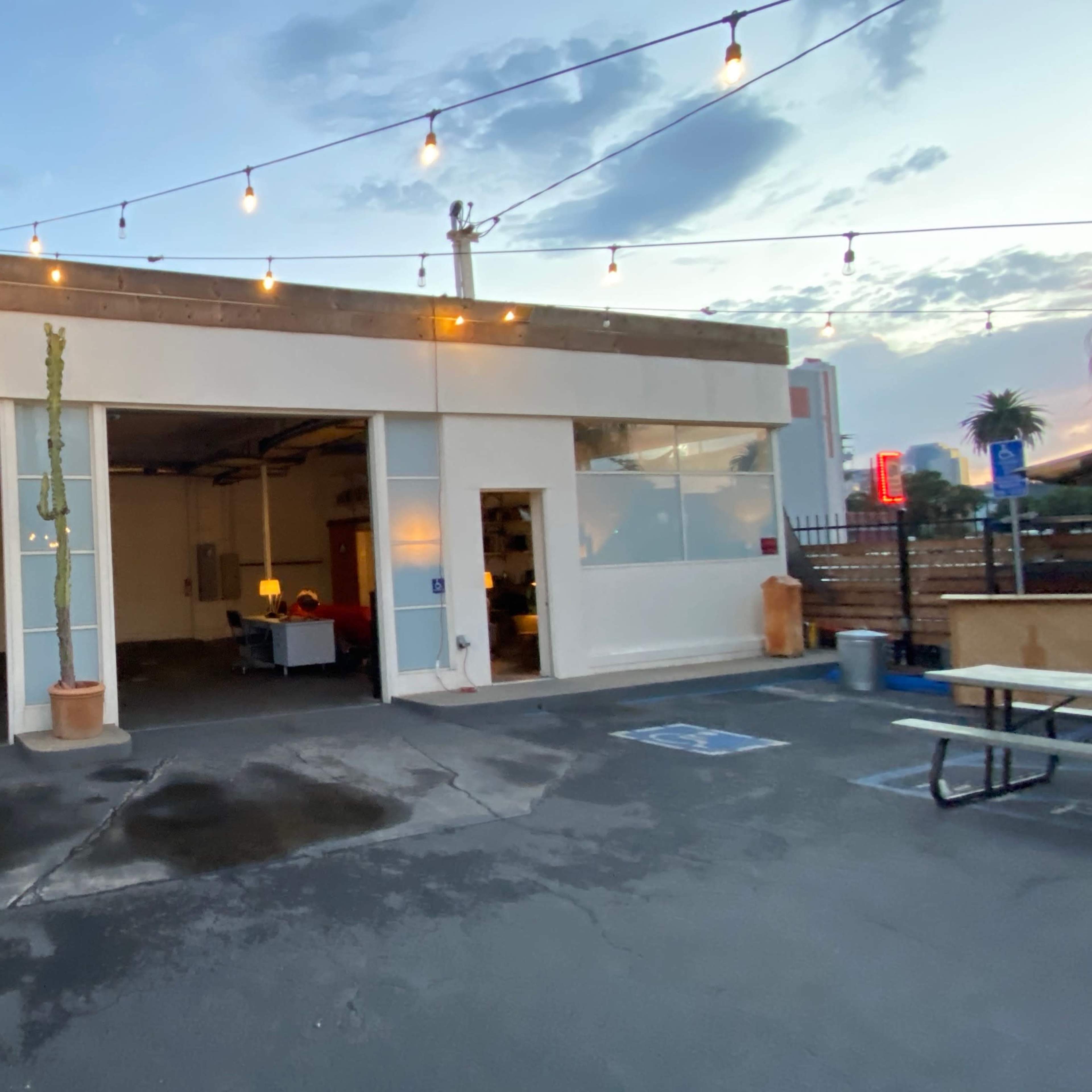 A partially enclosed industrial space with large windows, surrounded by string lights and featuring a few outdoor seating arrangements.