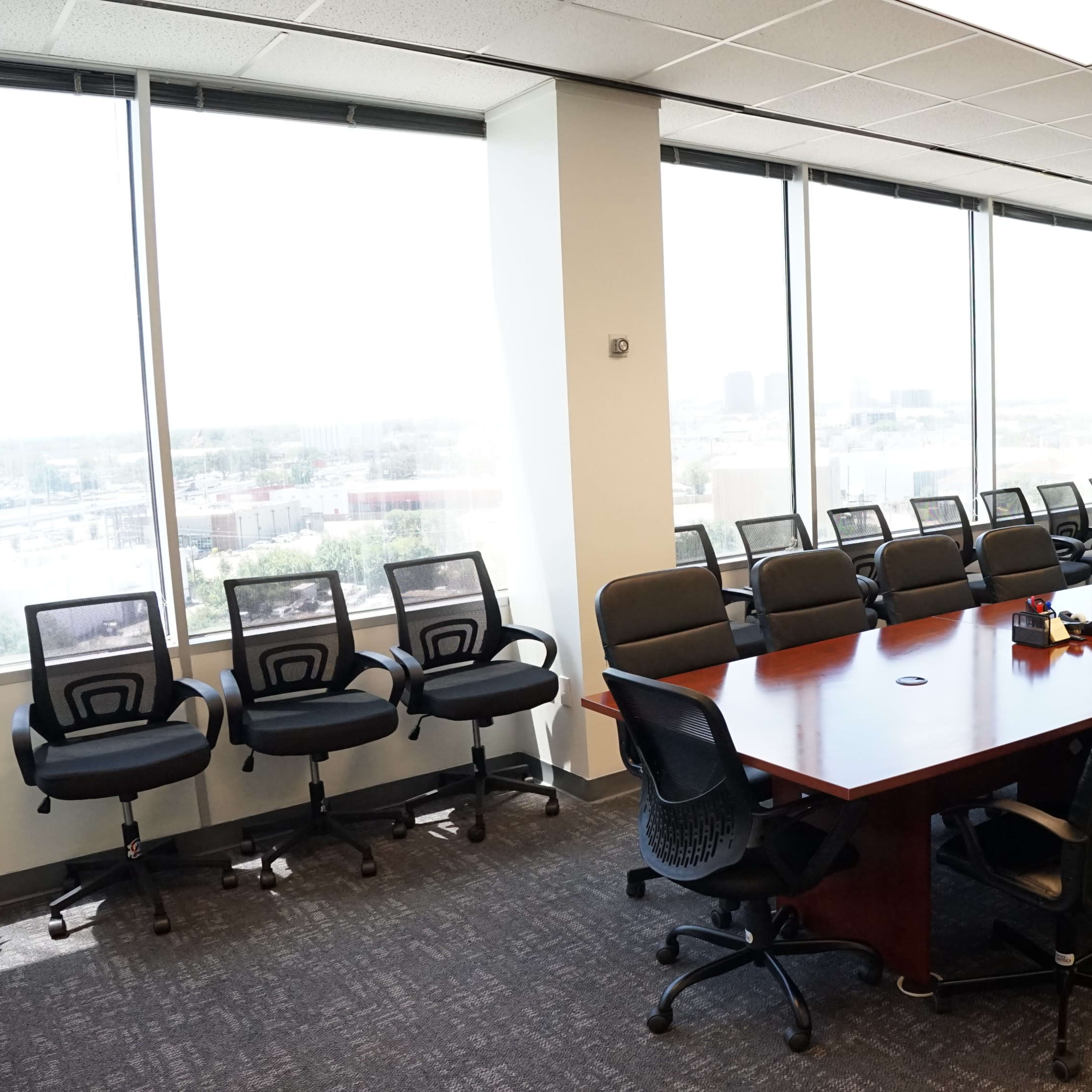 A conference room features a large wooden table surrounded by black office chairs, with floor-to-ceiling windows overlooking the city and a television mounted on the wall.
