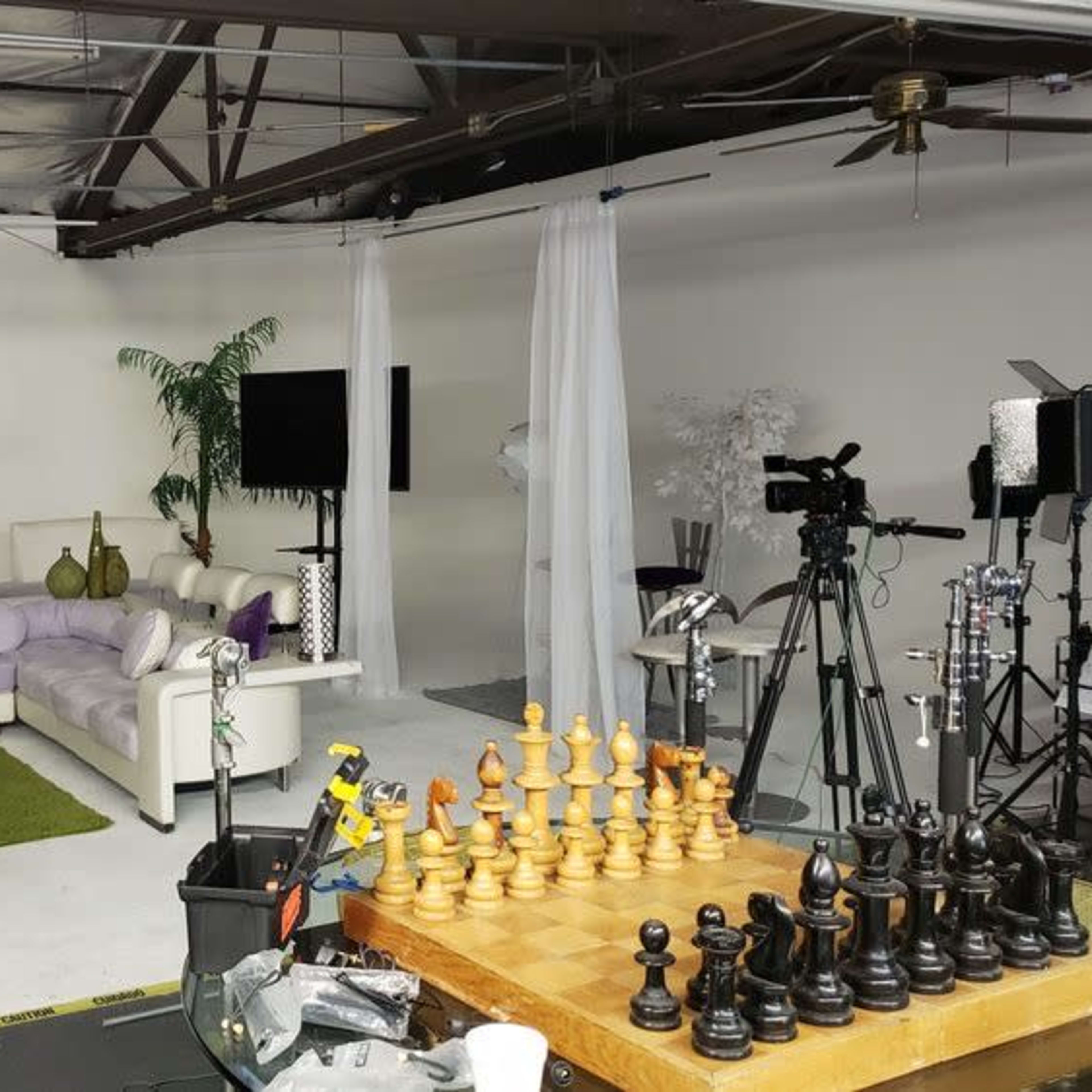A studio space featuring a white couch with purple accents, a chess set in the foreground, and various cameras and lighting equipment arranged around the room.