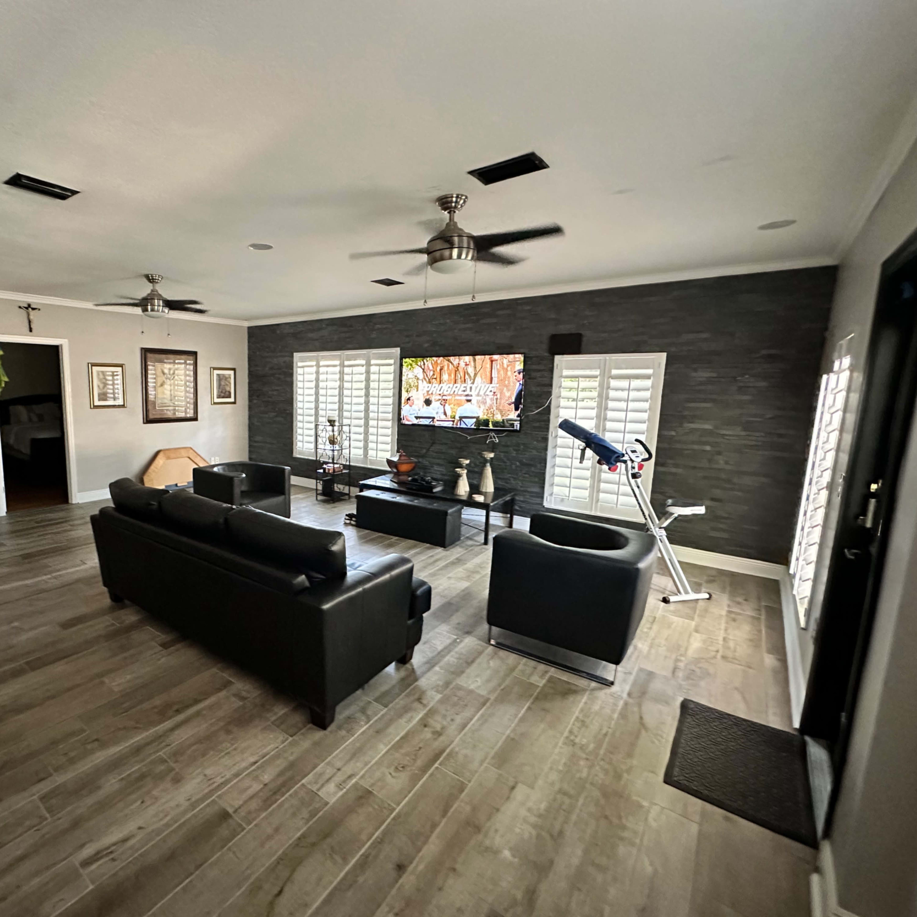 A spacious living room with dark leather furniture, a large flat-screen TV mounted on a stone wall, and exercise equipment in the corner.