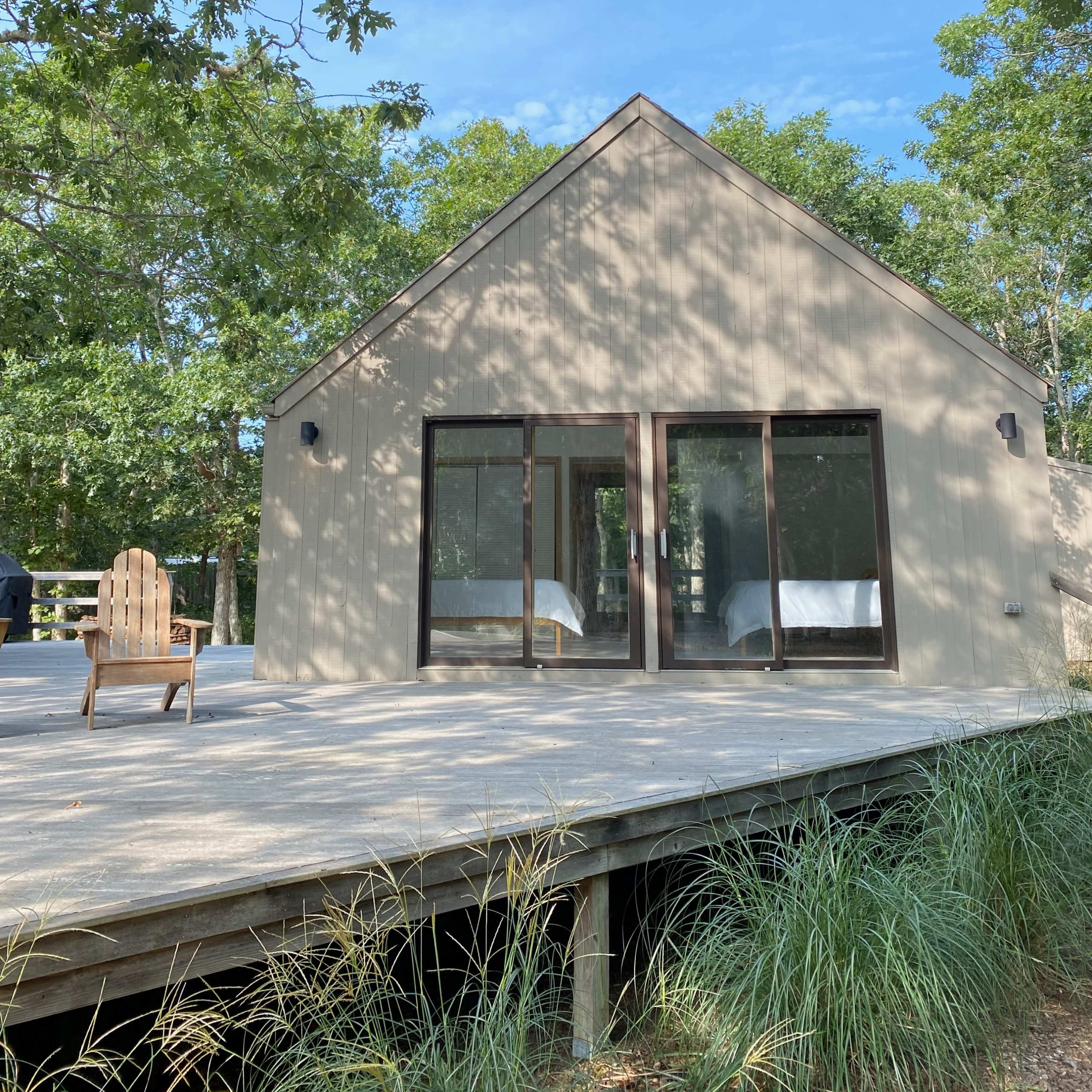 A modern, minimalist house with large windows is surrounded by trees and features a wooden deck with an Adirondack chair in the foreground.