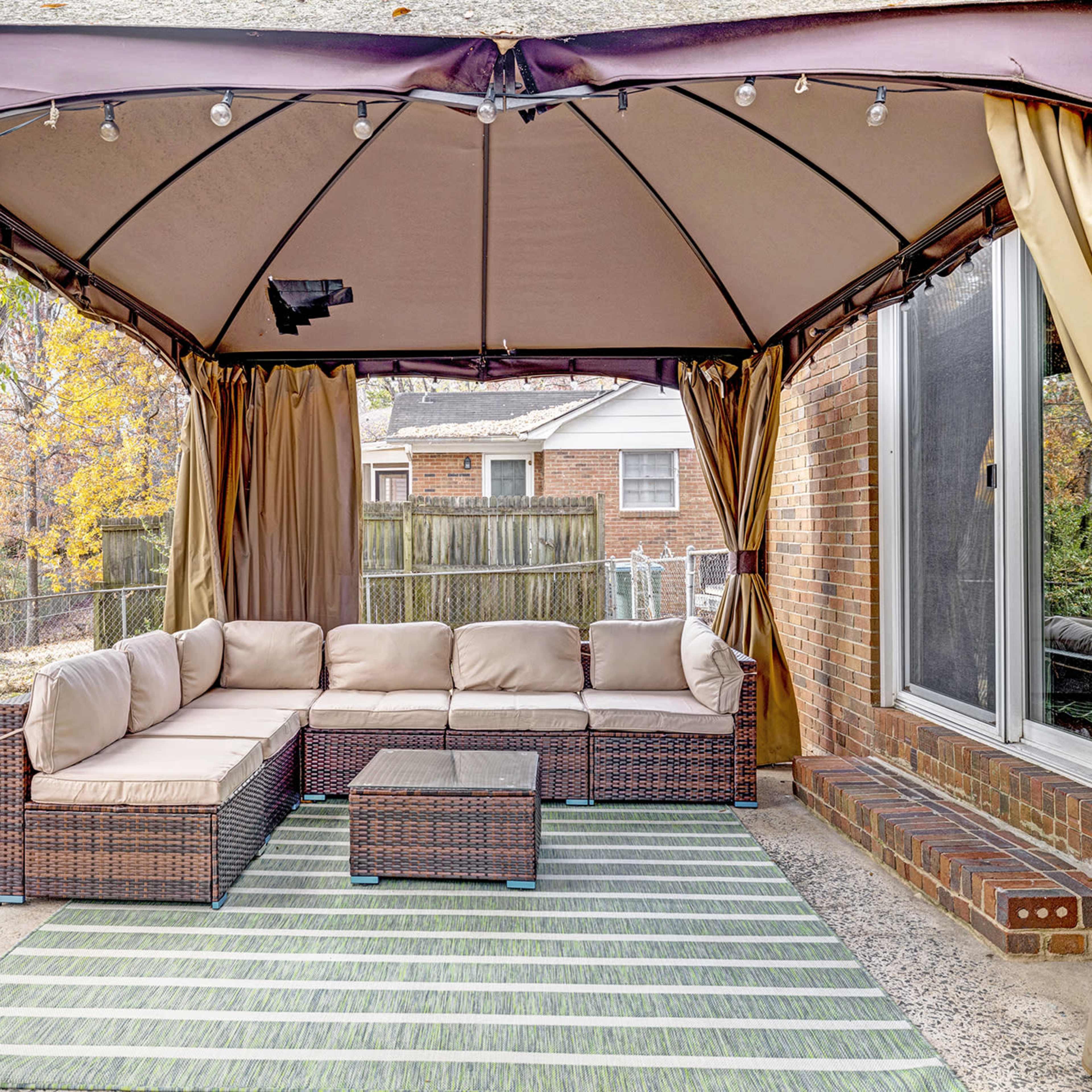 A covered patio area features a sectional sofa and a coffee table on a striped rug, with a backdrop of a fenced yard and neighboring houses.