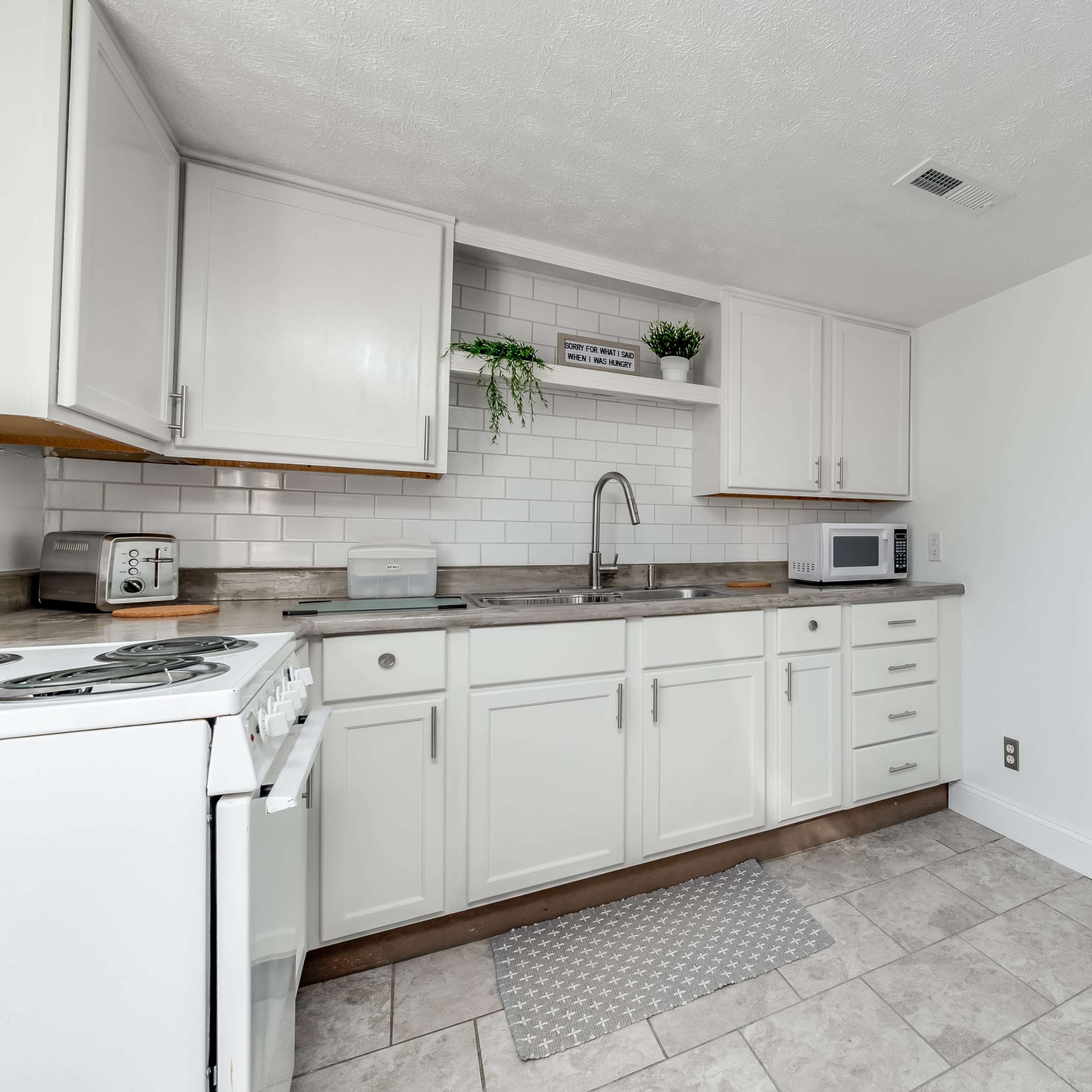 A clean kitchen with white cabinetry, a sink, a microwave, and tiled flooring, adjacent to a bathroom area.